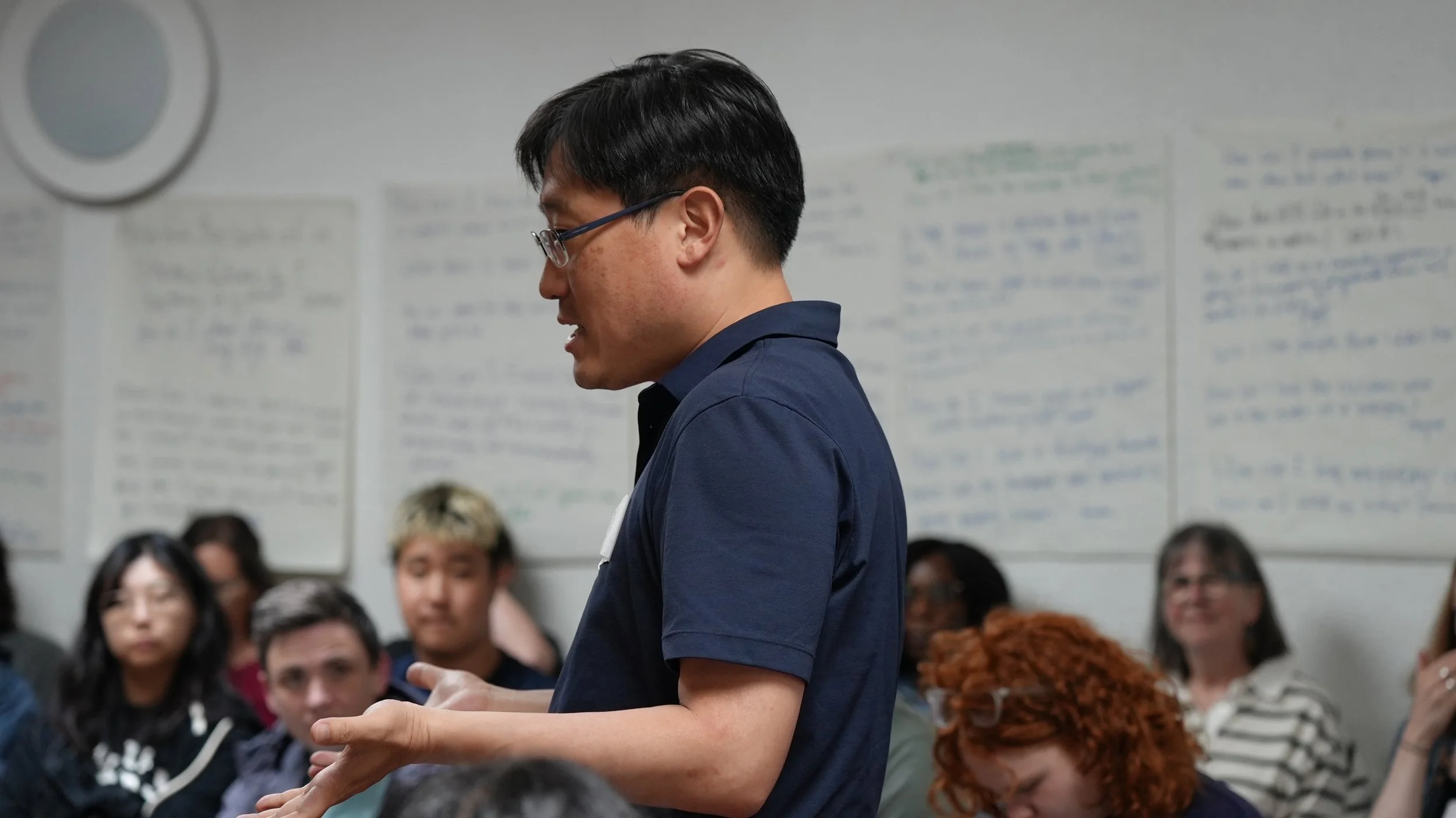 A man with glasses and black hair wearing a navy blue shirt speaks to a group of people in a room with white walls and handwritten posters.