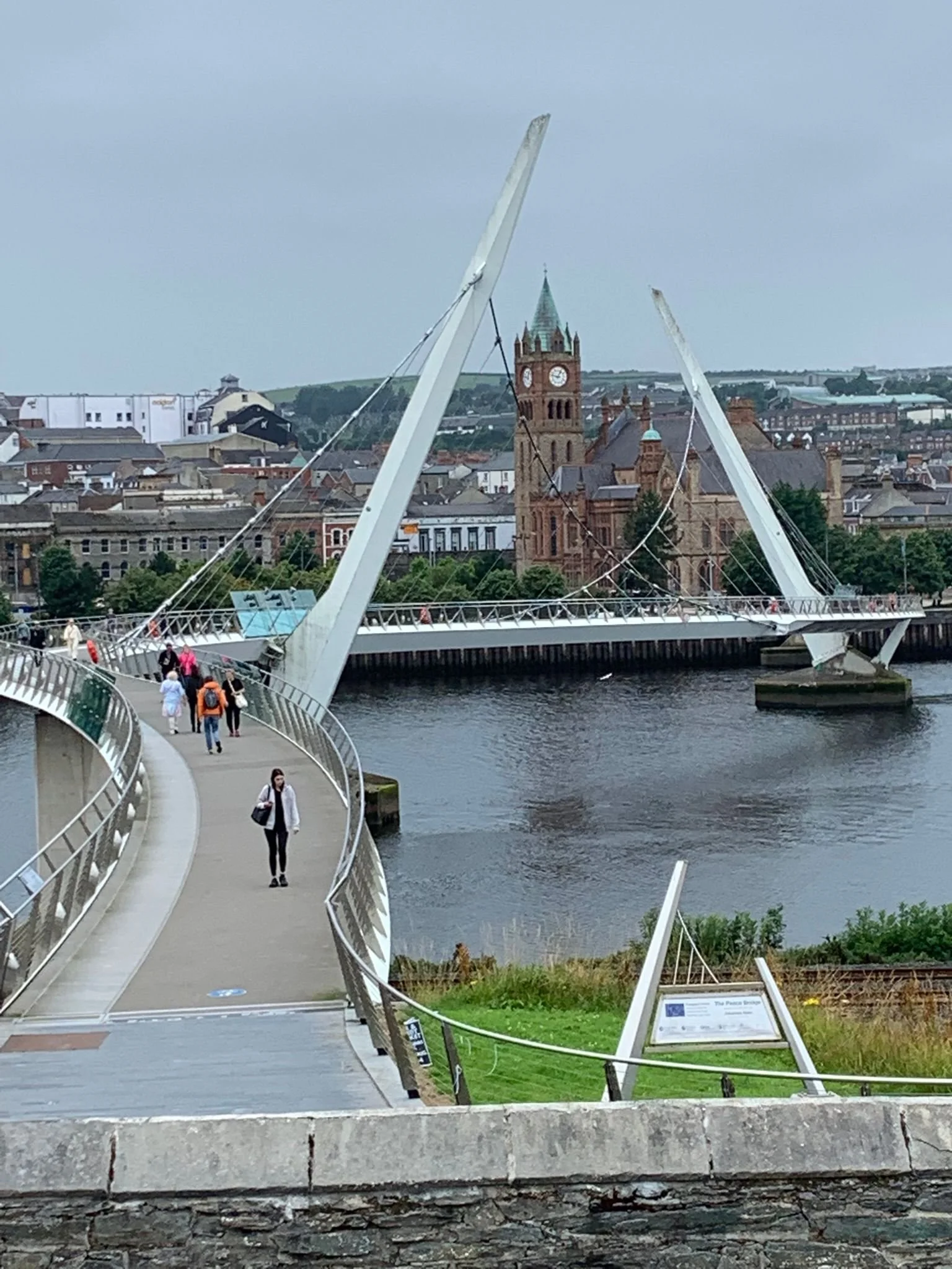 A pedestrian bridge over a river, with a cityscape and a historic red brick church with a clock tower in the background. The bridge features modern white support structures and a curved walkway with people walking.