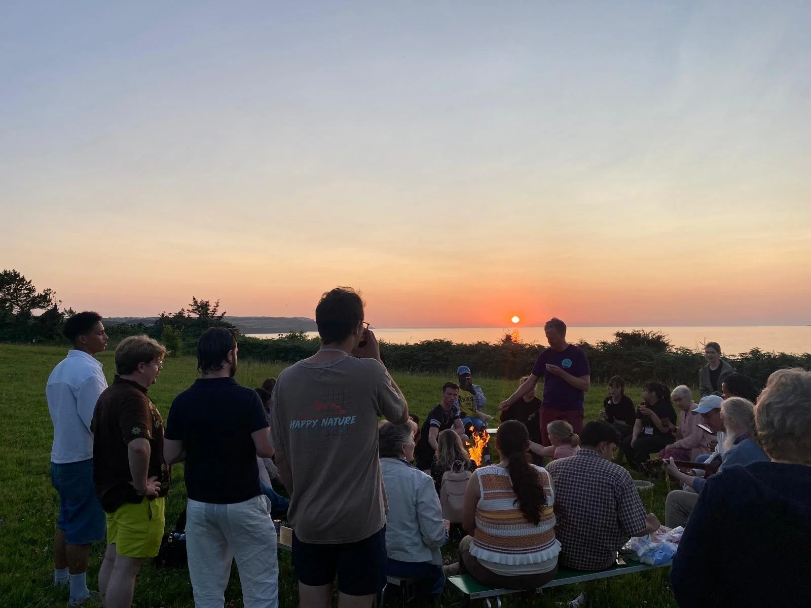 A group of people gathered outdoors on a grassy area at sunset, some standing and some sitting, with a view of the ocean and a setting sun in the background.