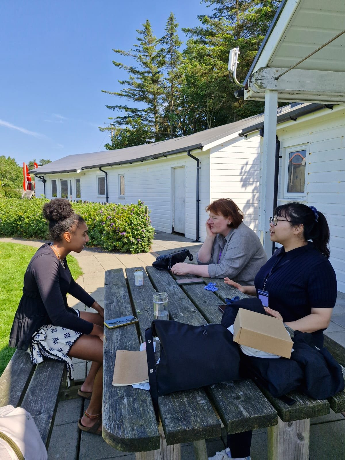 Three women sitting at a wooden picnic table outside, engaged in conversation on a sunny day, with a white building and green trees in the background.