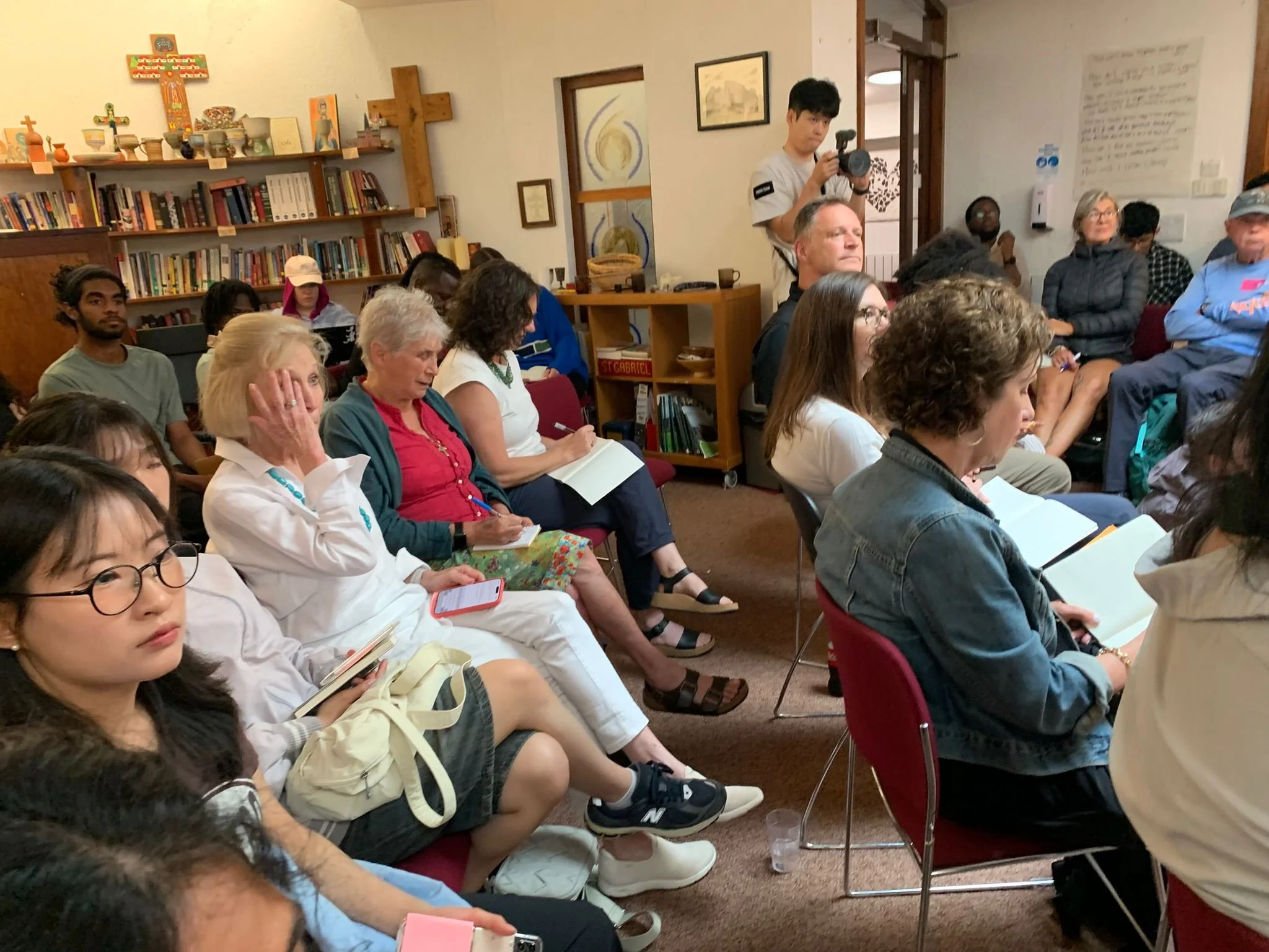 A group of people seated indoors, some taking notes or using electronic devices, attending a meeting or presentation, with a bookshelf, crosses, and religious images in the background.