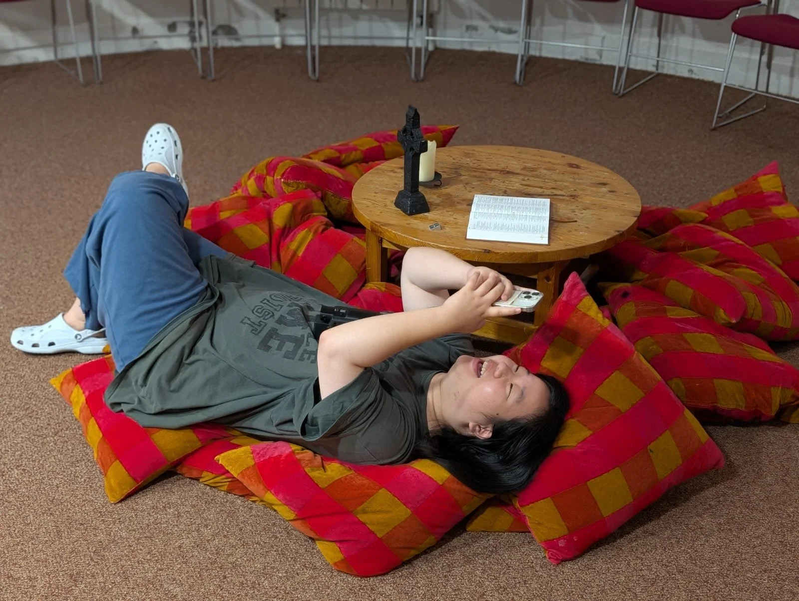 Person lying on the floor, relaxed on red and yellow checkered cushions, using a smartphone. A round wooden coffee table with a candle, cross, and open book is in front. Behind, a wall with chairs stacked.