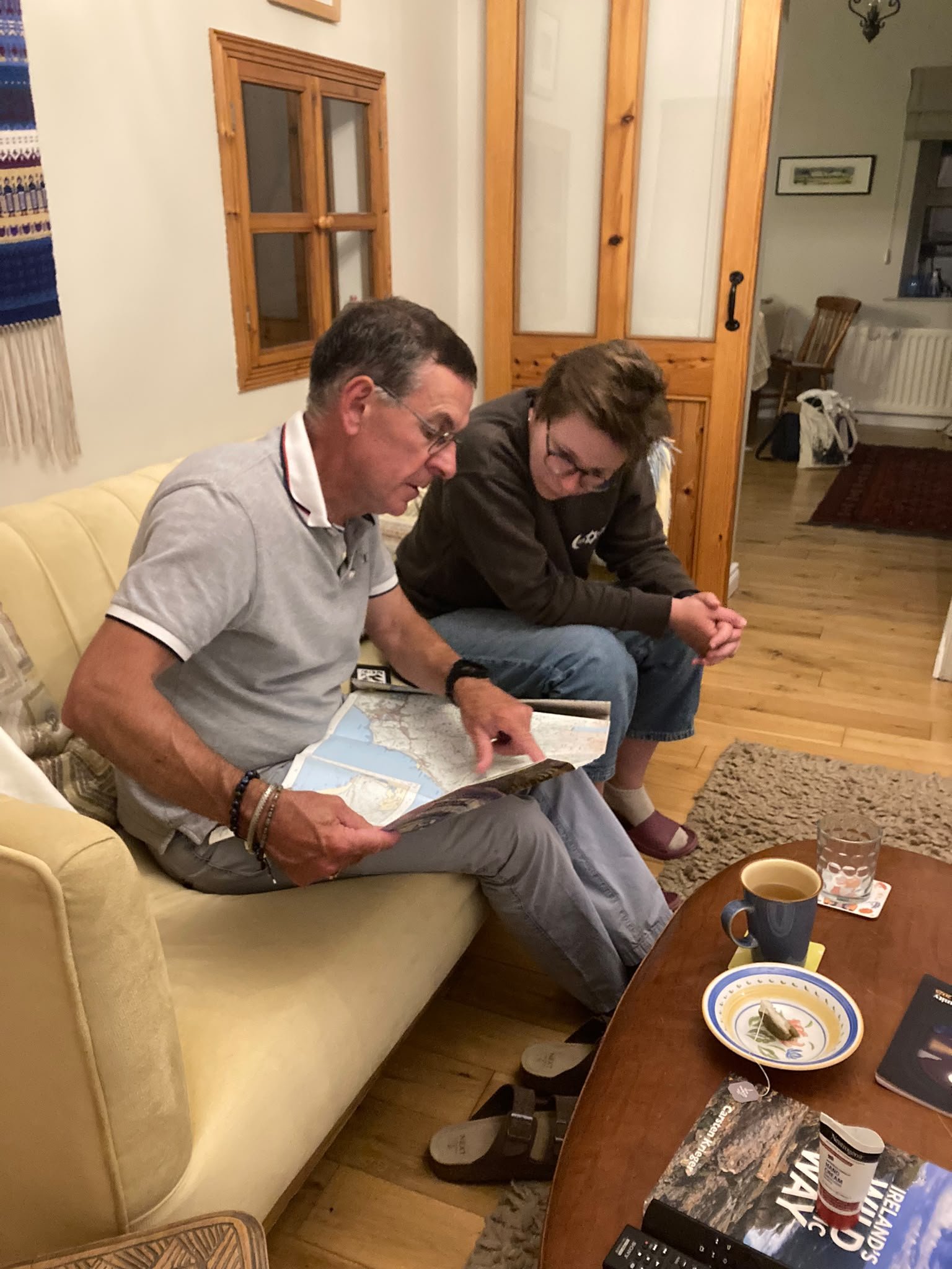 Two men sitting on a yellow couch, looking at a map together in a cozy living room with wooden accents, a wooden chair, some drinks on the table, and various household items in the background.