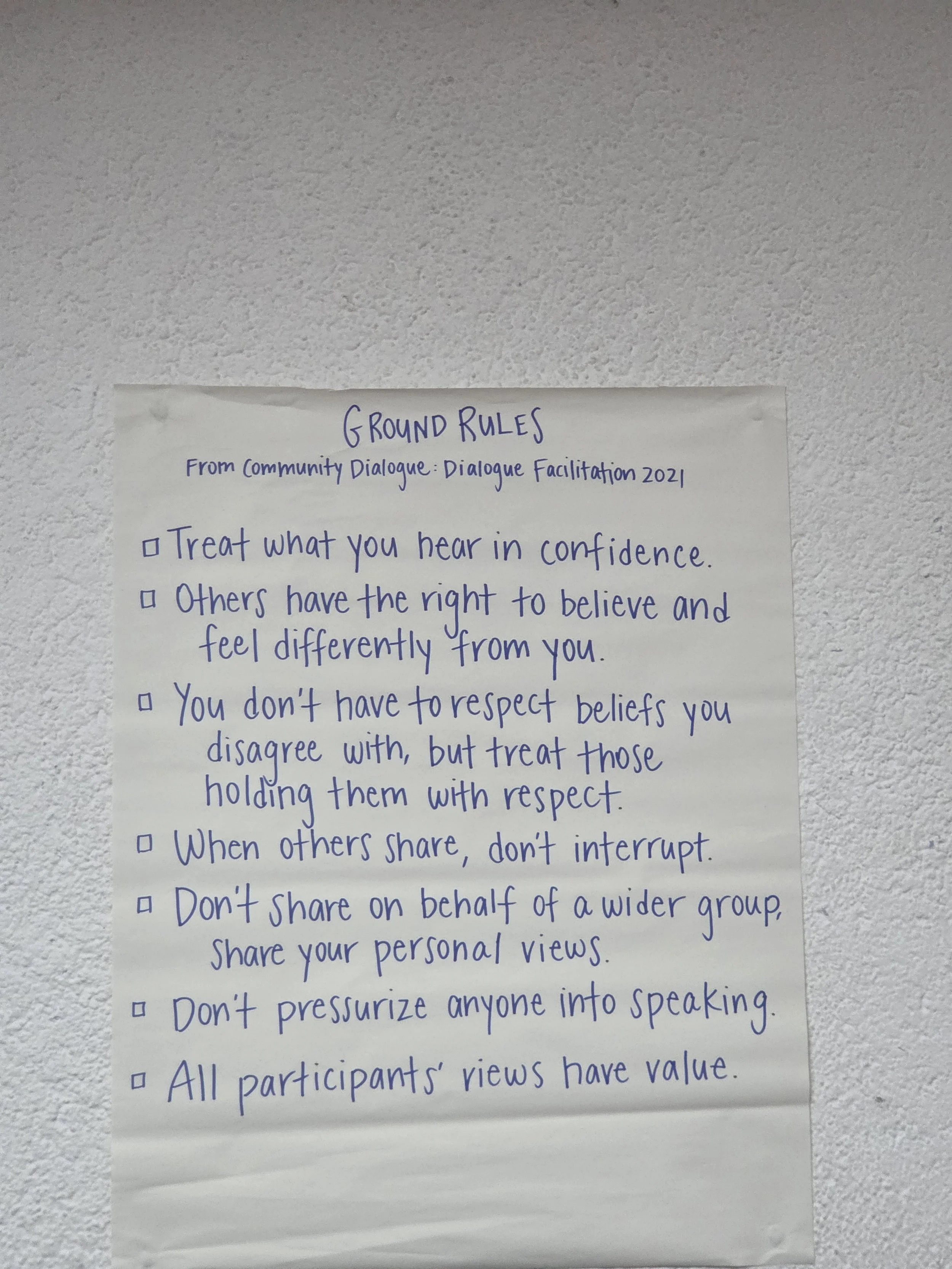 A handwritten list of ground rules for community dialogue on a piece of paper attached to a textured white wall.