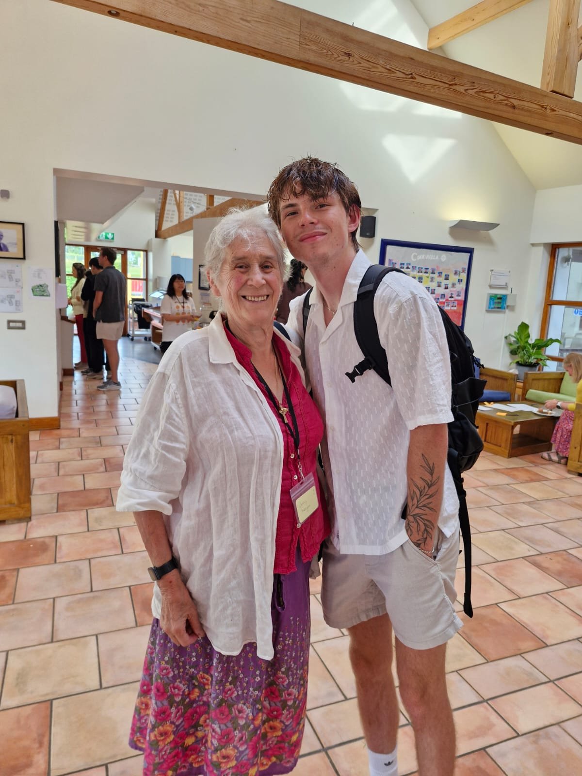 A smiling older woman and a young man with a tattoo on his arm standing inside a building with wooden beams and tiled floor, posing for the photo.