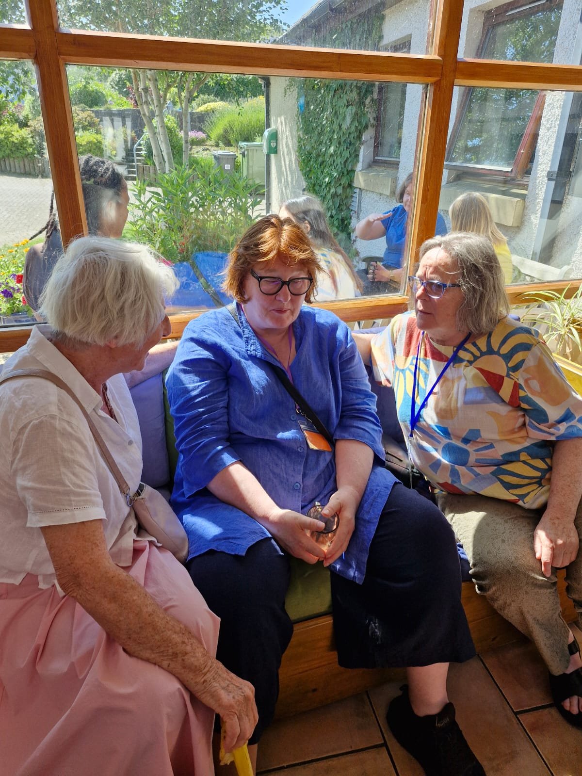 Four women are sitting together inside a sunlit room with large windows, having a conversation. Outside, a garden with trees and flowers is visible.