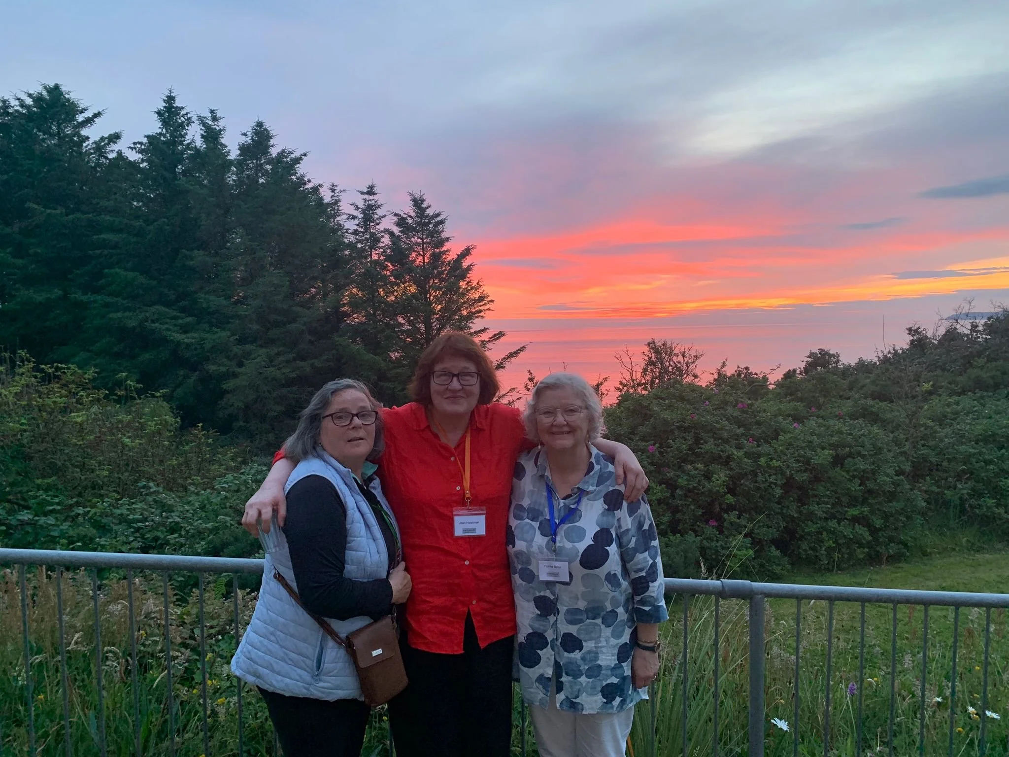 Three women standing together outdoors during sunset with a background of trees and a colorful sky, smiling and posing for the photo.