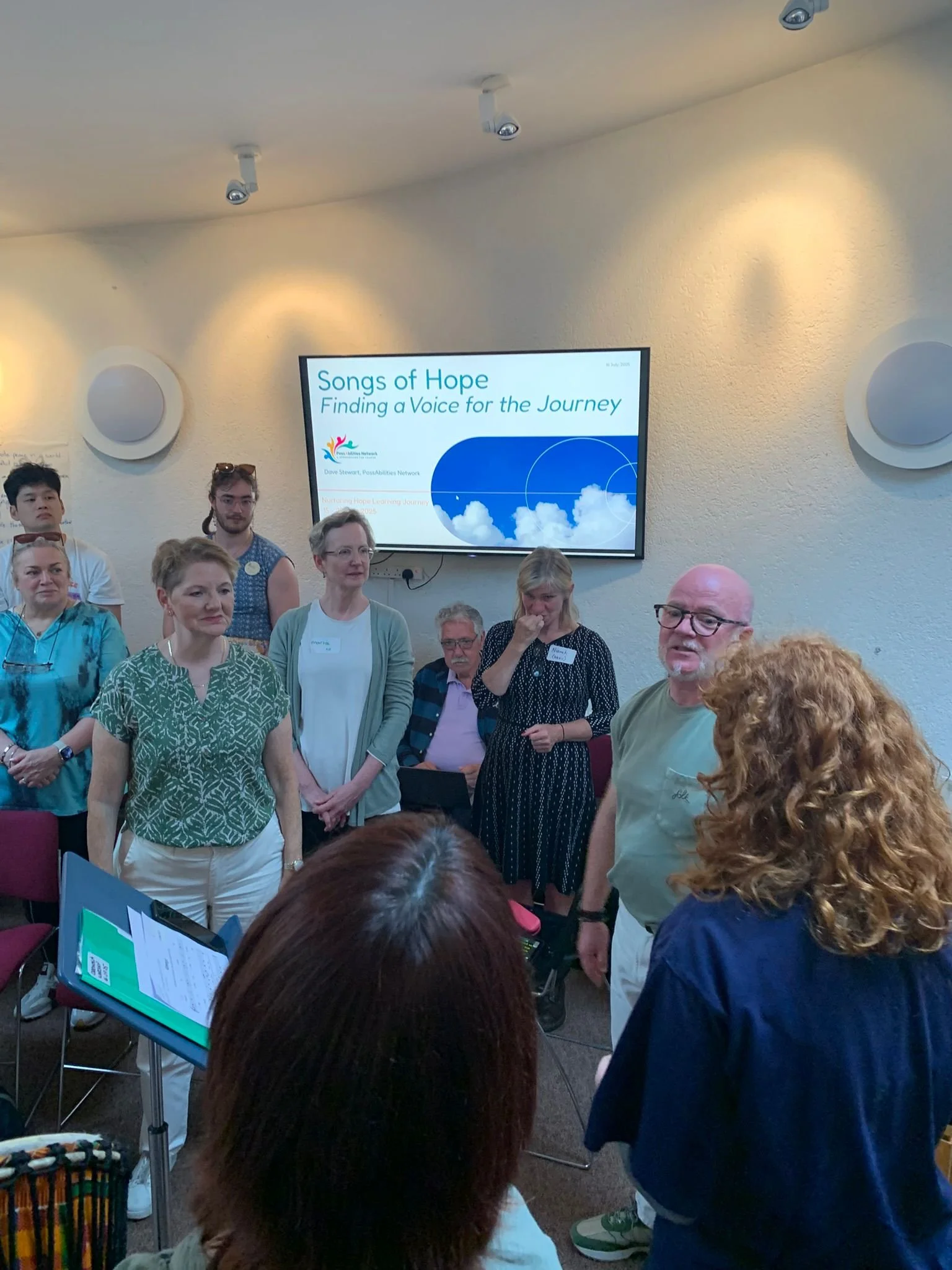 Group of people attending a presentation titled 'Songs of Hope: Finding a Voice for the Journey' in a conference room with a large screen and some speakers on the wall.
