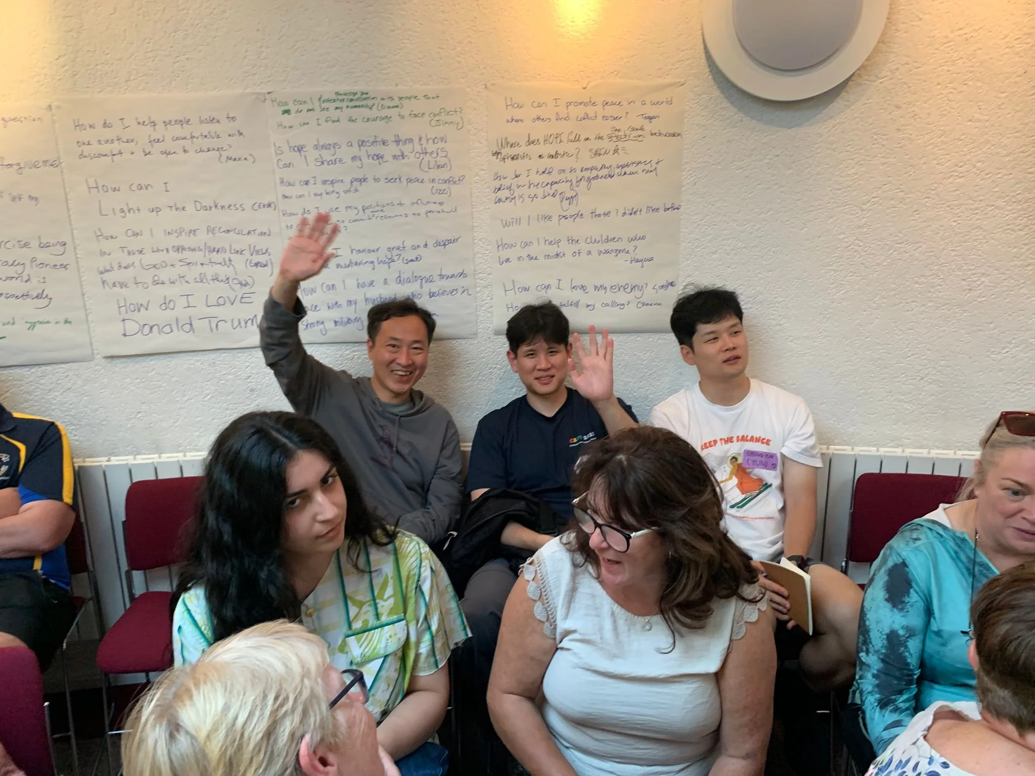 Group of diverse people sitting and standing in a room, some smiling and waving, with handwritten posters on the wall behind them.