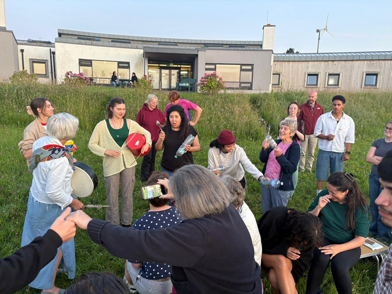 A group of people gathered outdoors on a grassy hill, engaging in storytelling or a communal activity, with some holding water bottles and one person playing a drum, in front of a modern building and wind turbine.