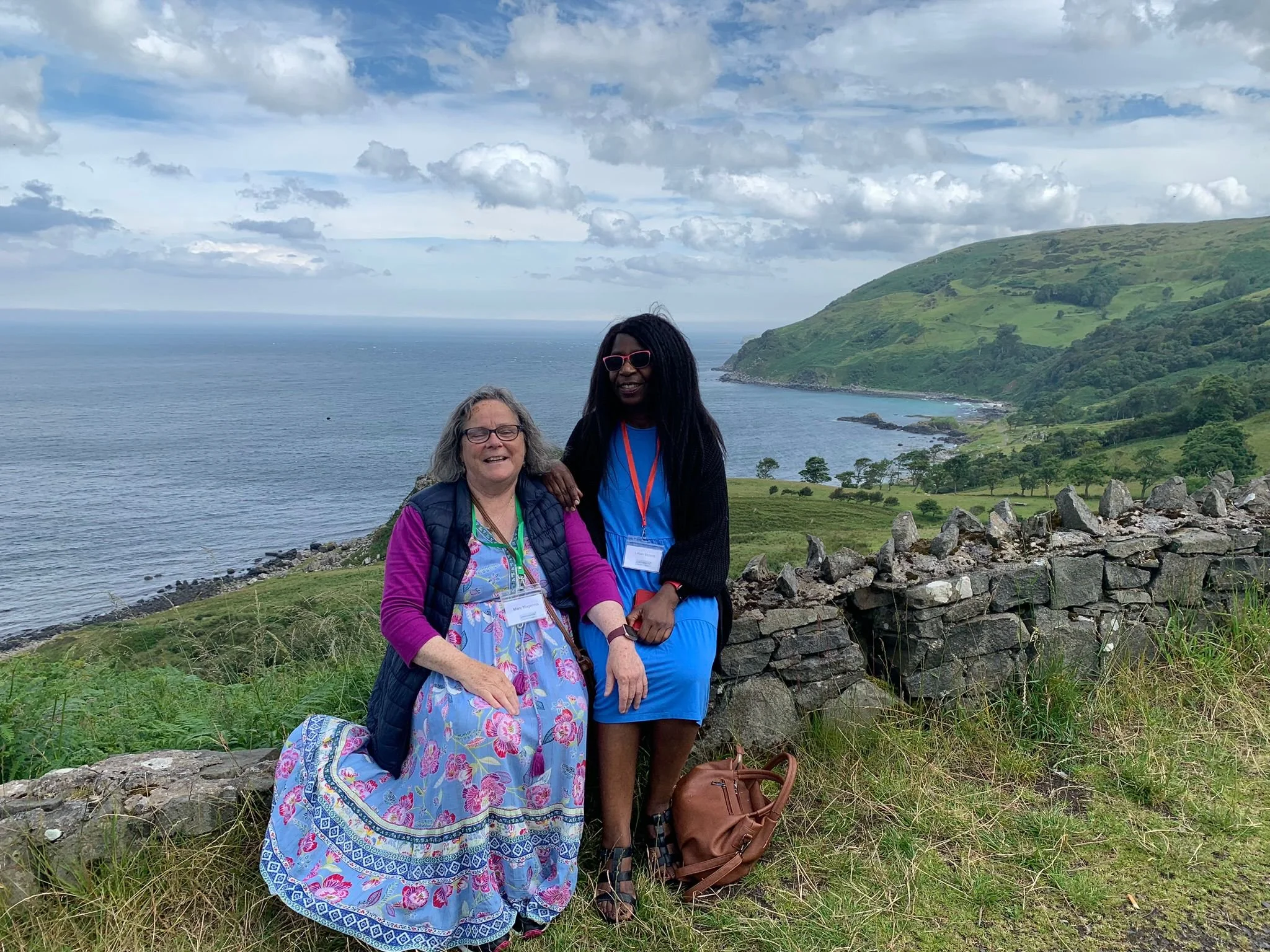 Two women standing outdoors near a stone wall with a scenic coastal landscape, grassy hills, and the ocean in the background.