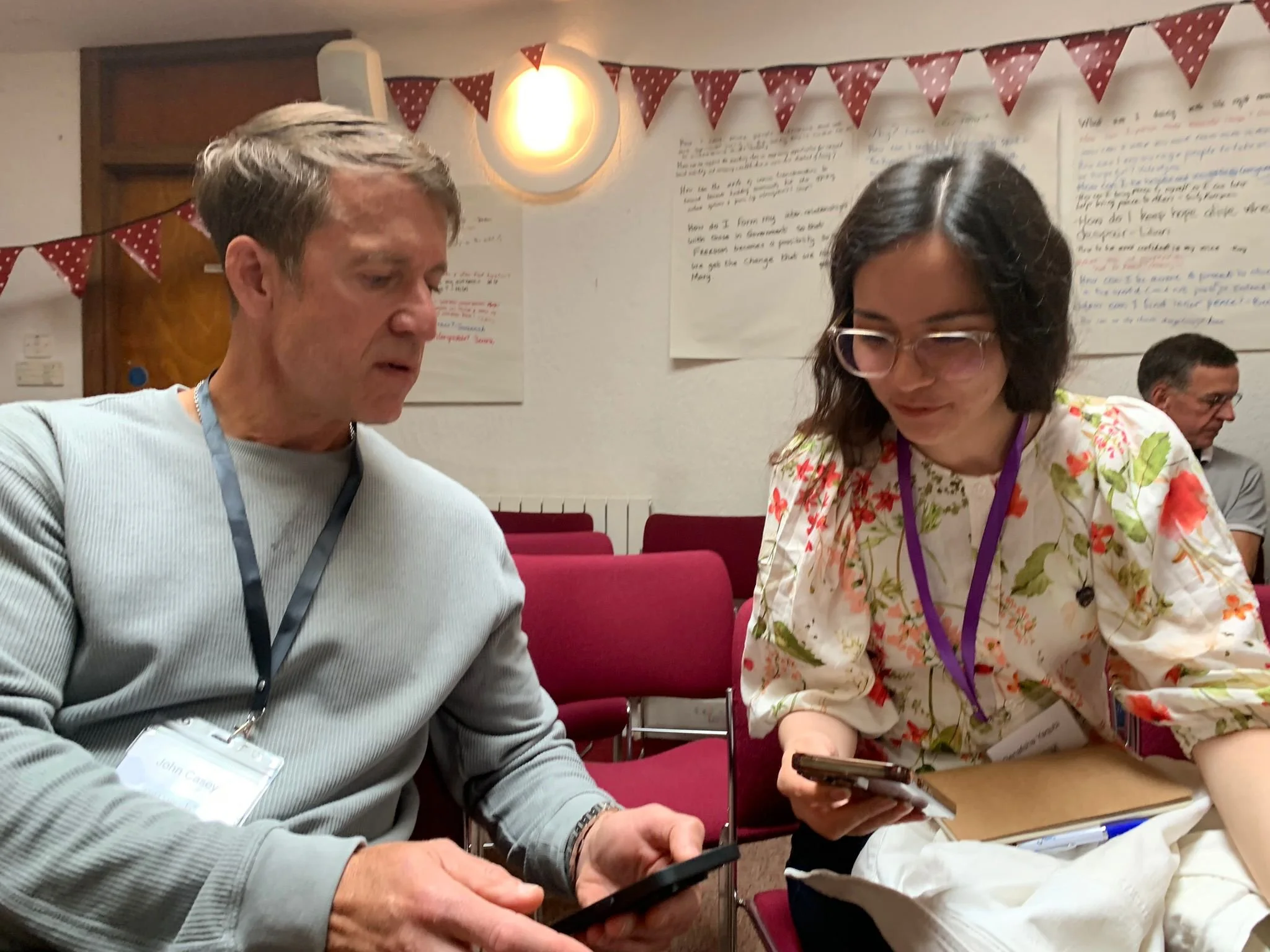 Two people sitting at a table looking at their mobile phones in a conference or meeting room with red chairs and handwritten posters on the wall.