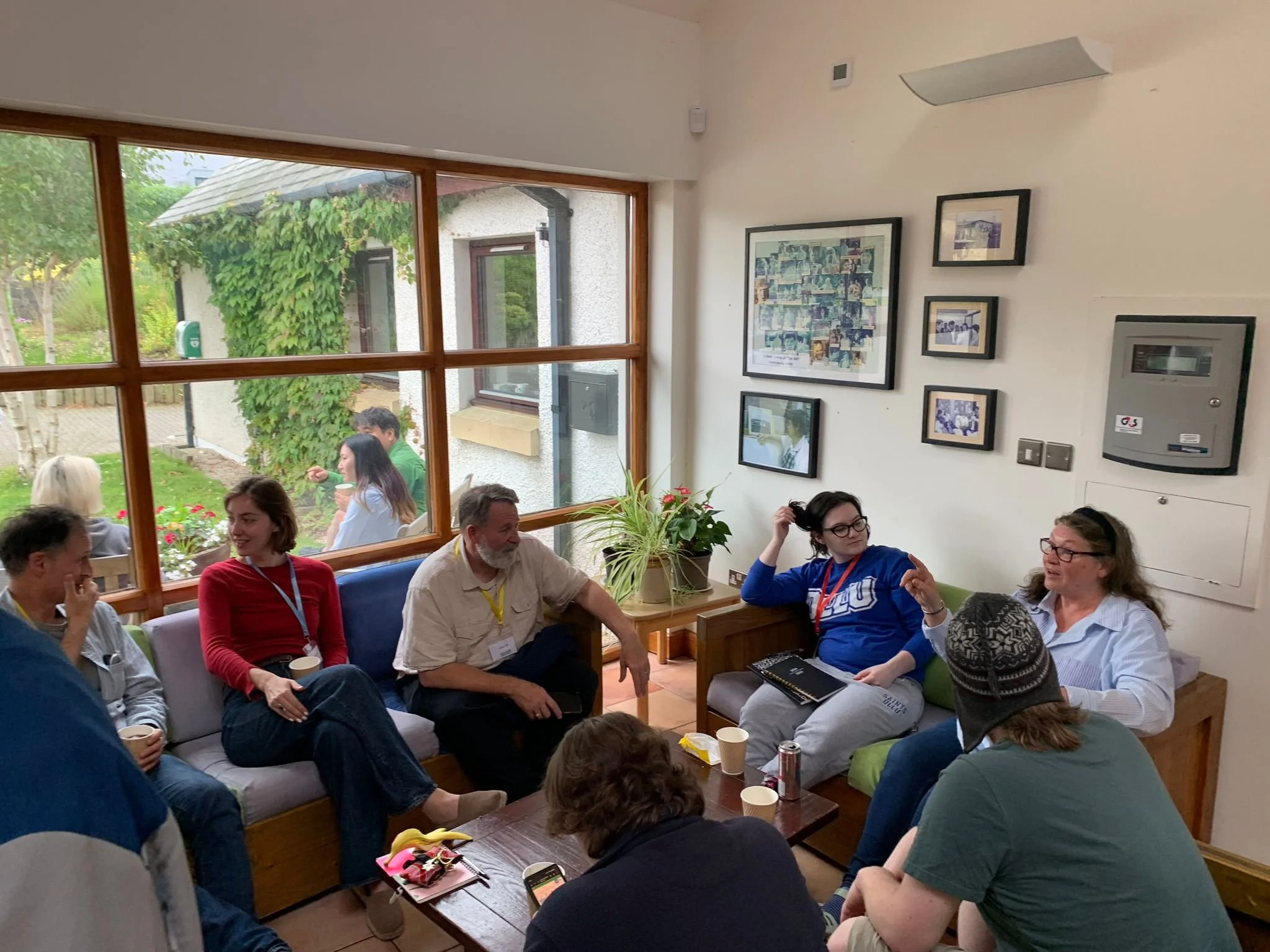A group of people sitting indoors and outside on a patio, engaging in conversation, with a large window showing a garden scene.