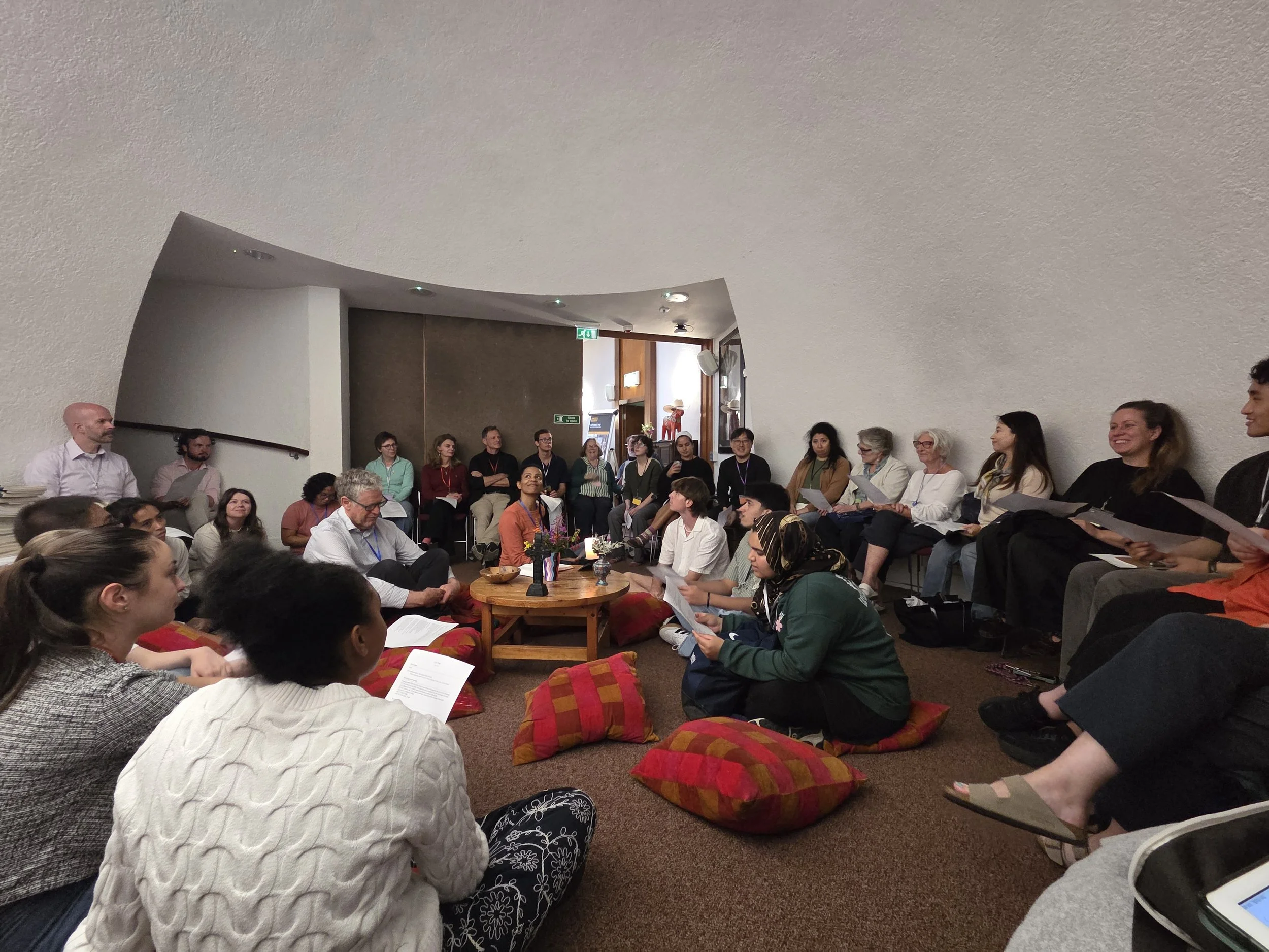 People are seated on the floor in a semi-circle in a room, attending a gathering or workshop. Some are reading papers, and there are cushions for sitting. The background shows additional attendees standing and sitting near a wall with artwork.