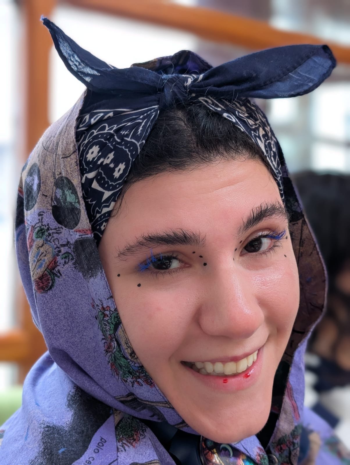 Close-up of a smiling young woman with dark hair, wearing a blue patterned headscarf and makeup with blue eyeliner and black dots around her eyes.