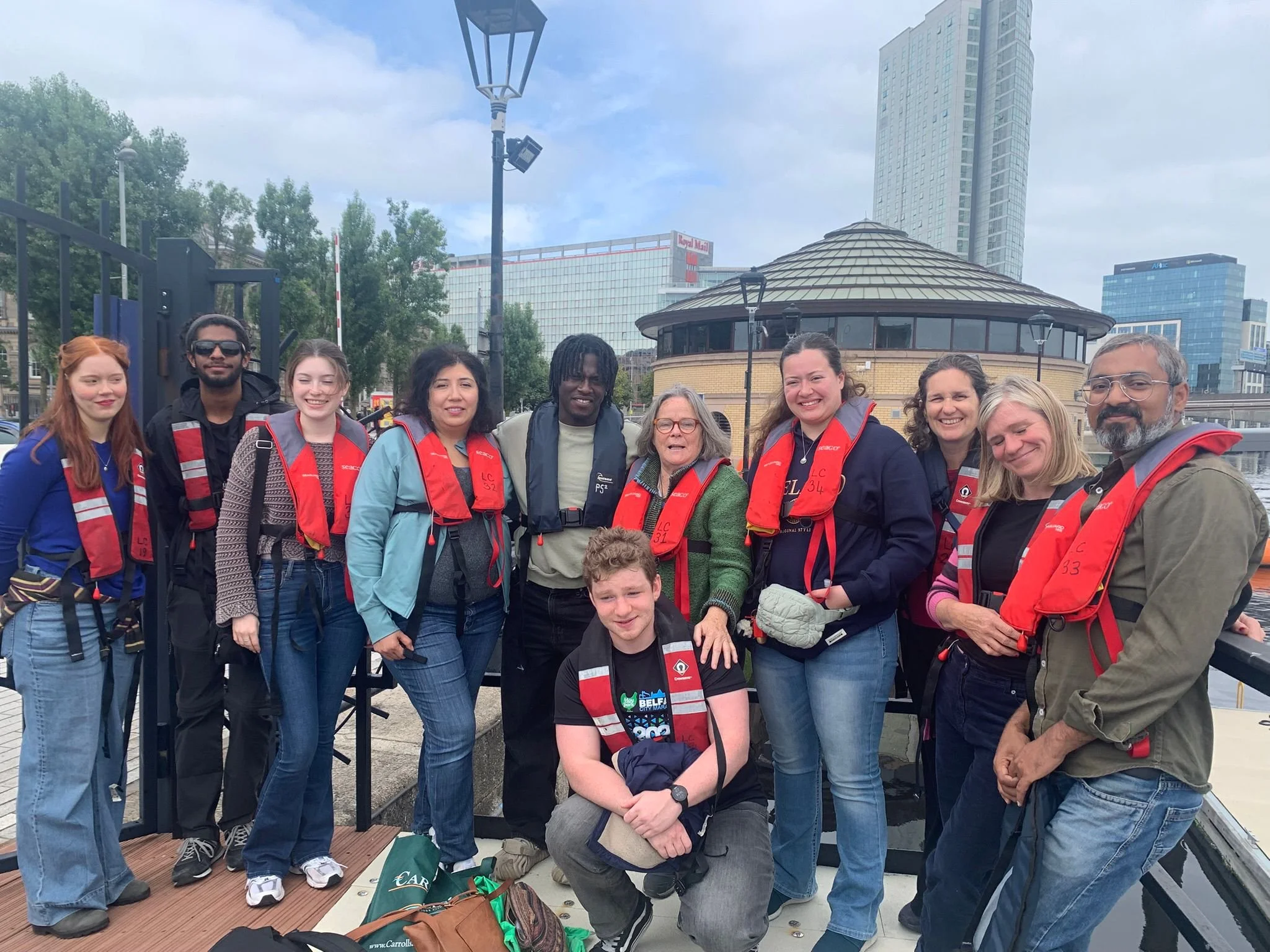 A group of eleven people standing outdoors in front of a cityscape, wearing red and gray life jackets and smiling at the camera.