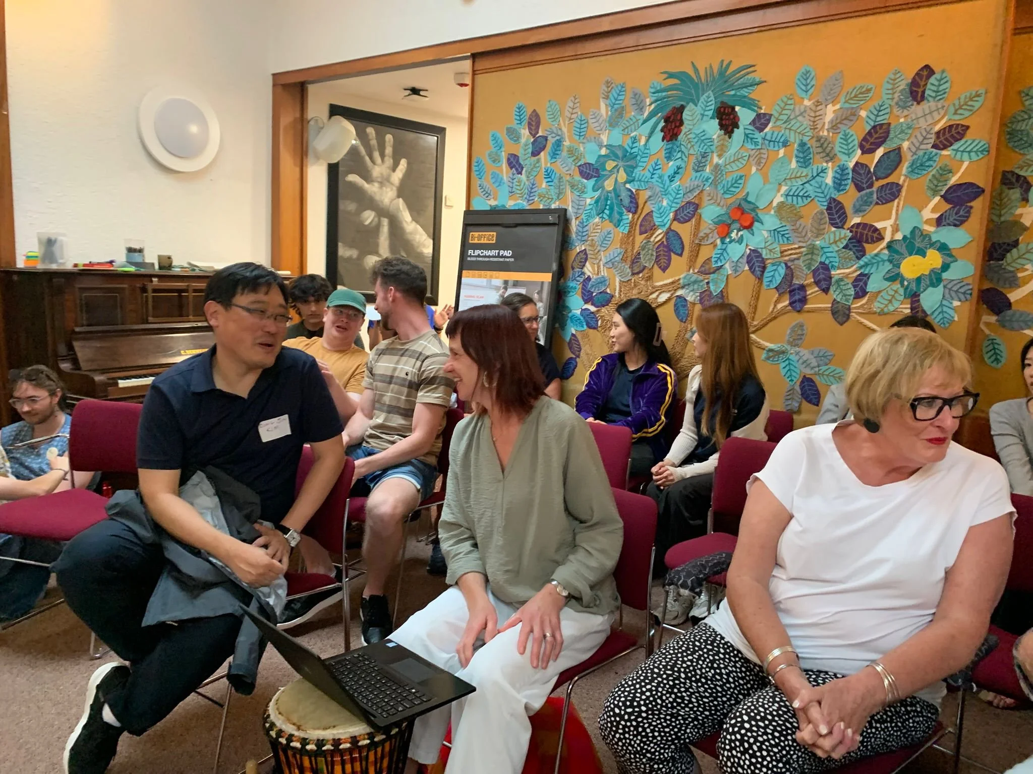 Group of people seated in chairs at an indoor event, engaging in conversations, with a colorful mural of leaves and berries on the wall behind them.