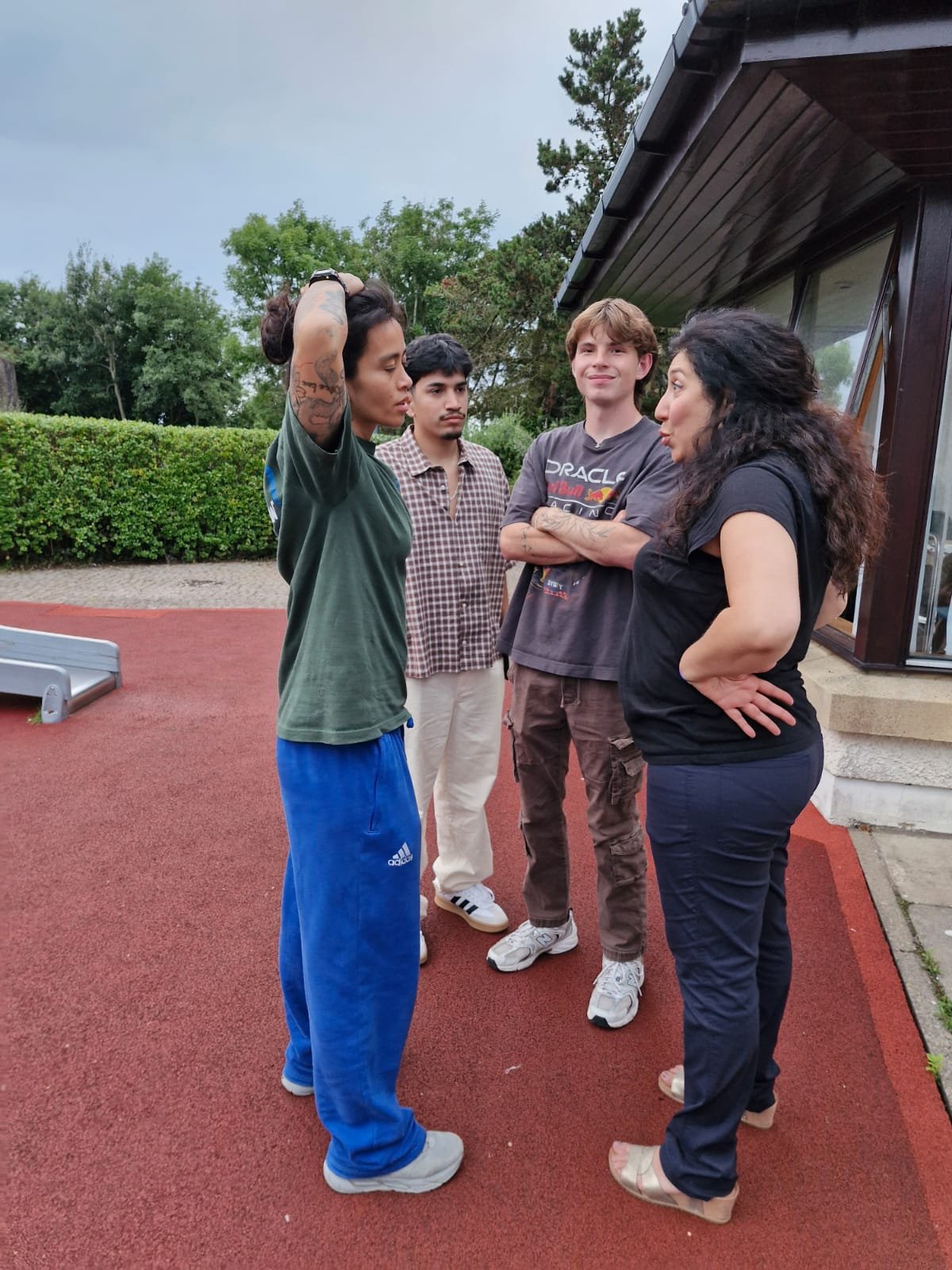 Four people standing on a red outdoor surface, engaged in a conversation, with greenery and a building in the background.