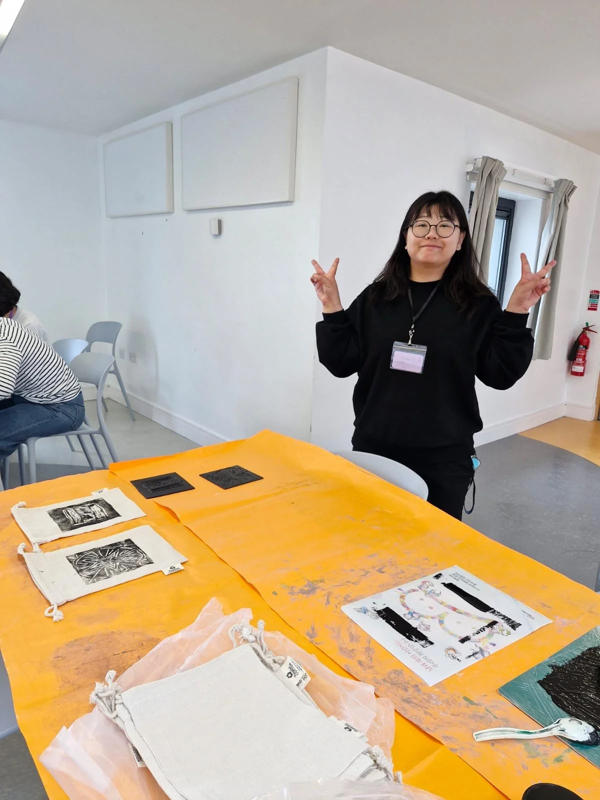 A woman with glasses and a black sweater dressed in black is making a peace sign with both hands in a bright white room. She has a name tag on a lanyard around her neck. There are printed materials and fabric bags on an orange table in front of her, 