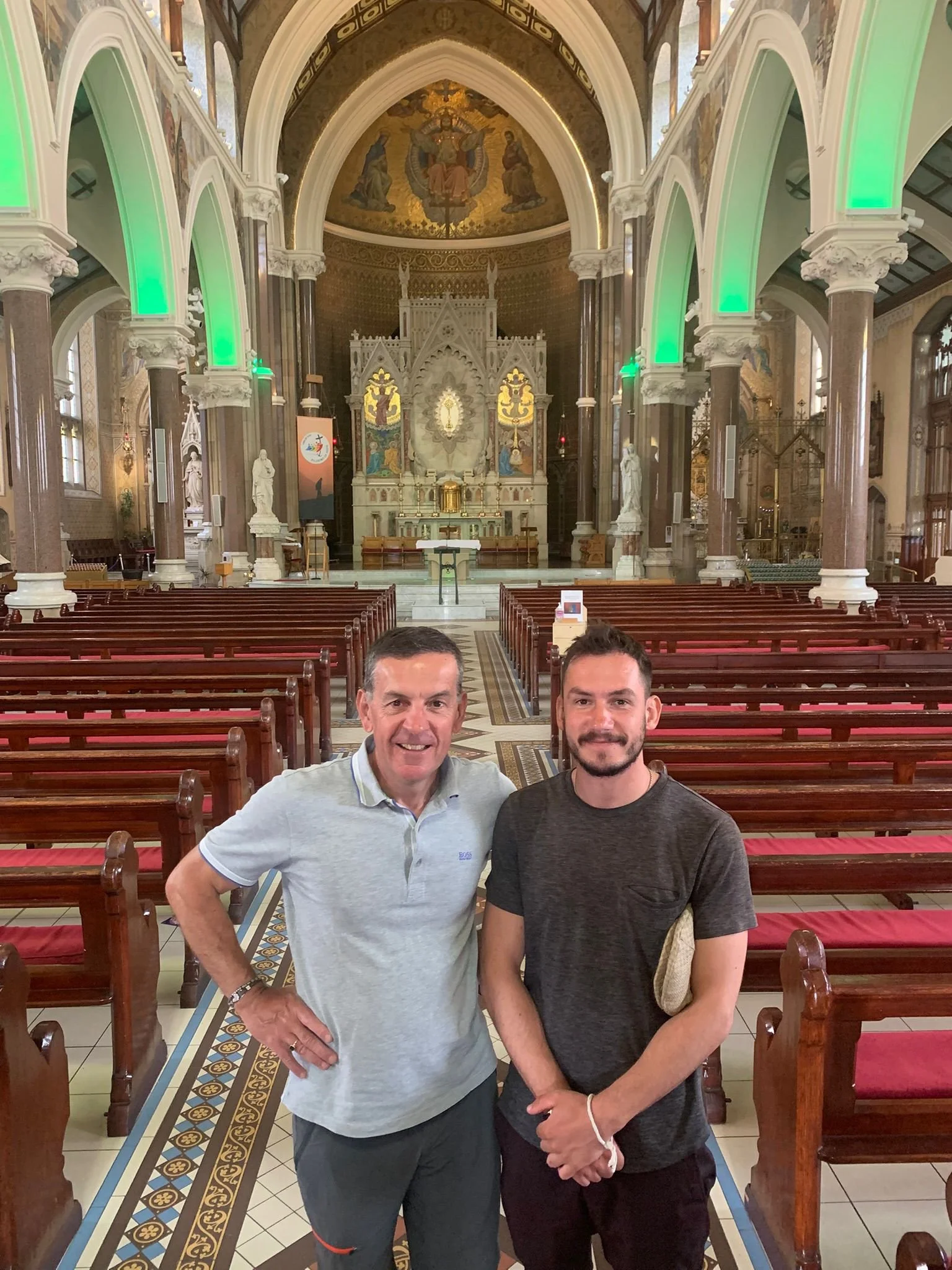 Two men standing inside a church, smiling. The church features high ceilings, ornate decorations, and wooden pews. The altar is visible in the background, illuminated by green lights on the arches.