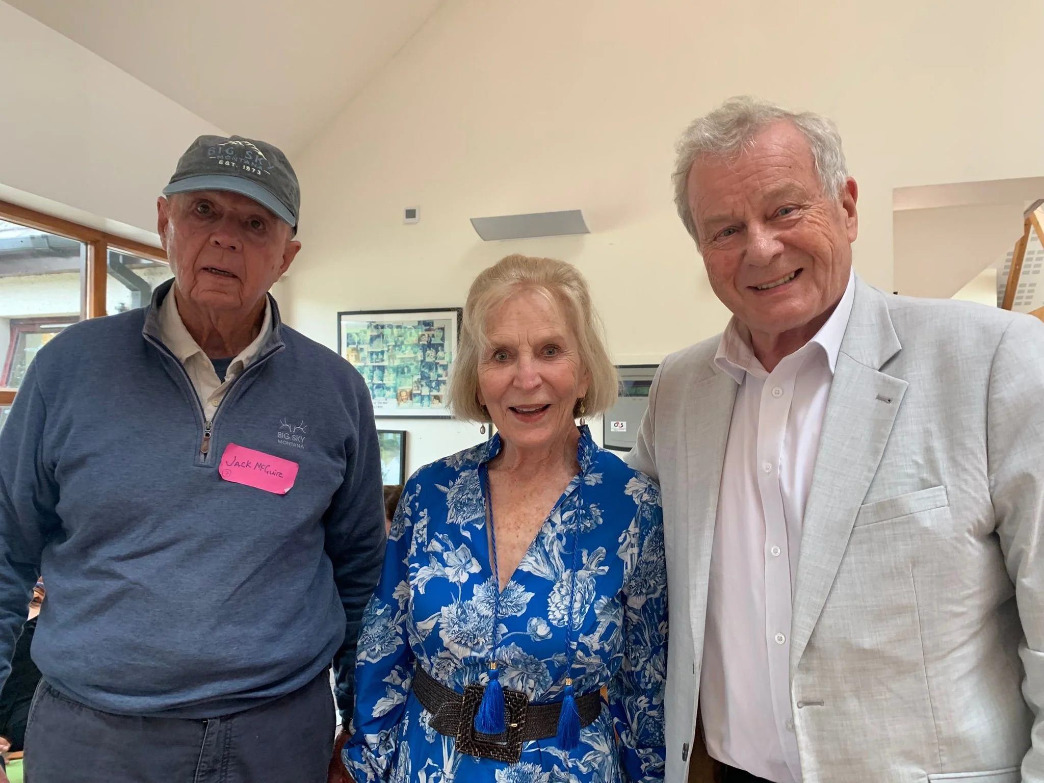 Three older adults standing indoors, smiling at the camera. The man on the left wears a gray cap and a blue zip-up jacket with a pink name tag. The woman in the middle has short blonde hair and wears a blue floral dress with a black belt. The man on 