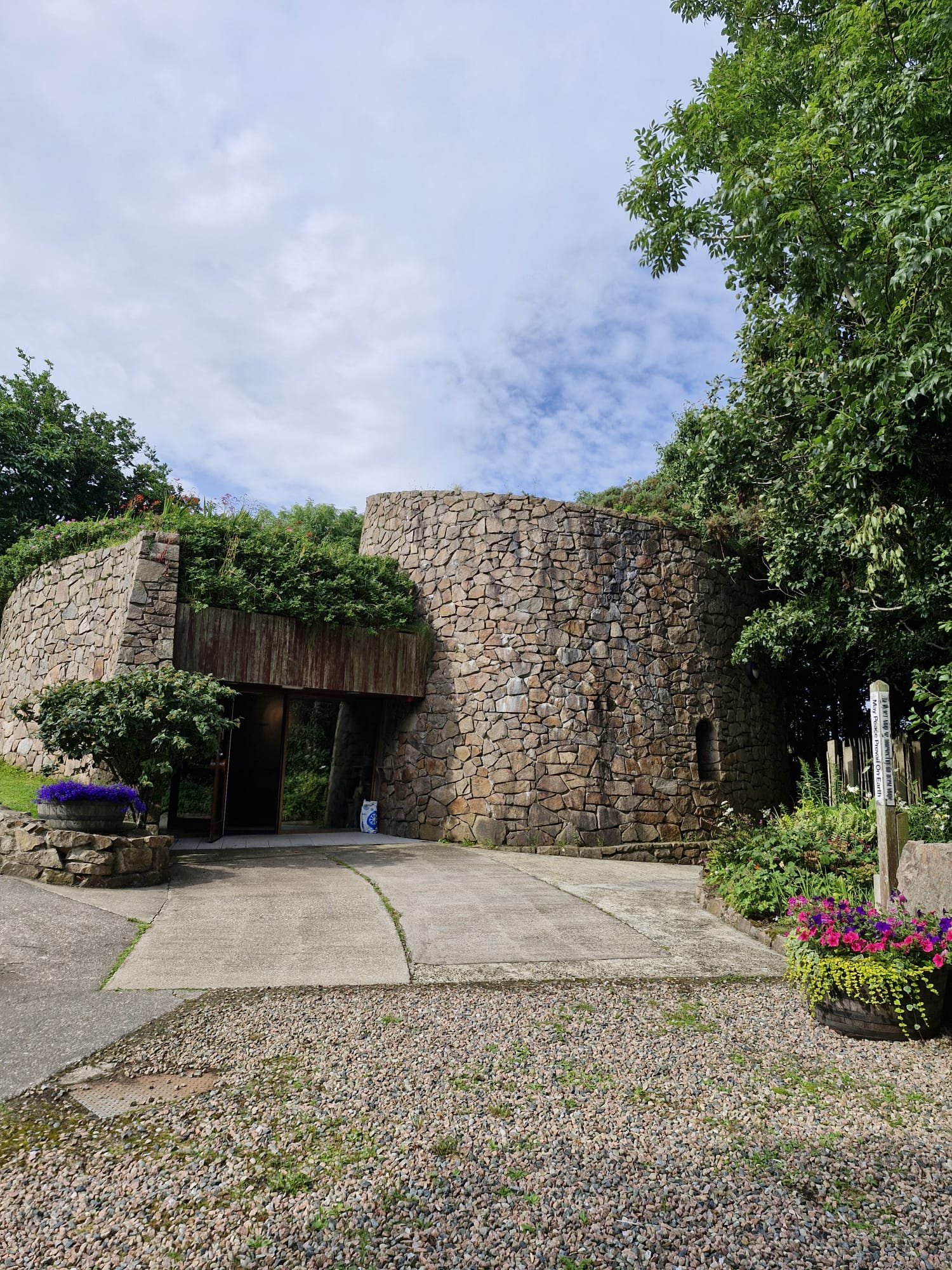 A stone building with a curved wall and large glass door, surrounded by greenery and colorful flowers on either side, under a partly cloudy sky.