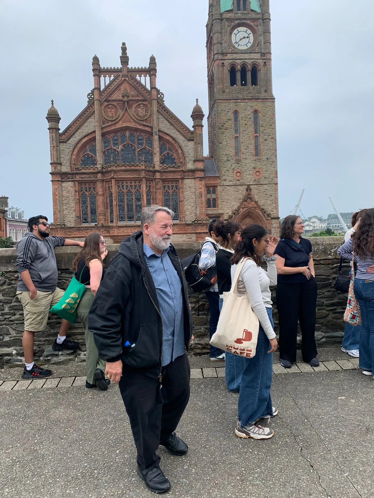 People standing outdoors near a historic stone church with a tall clock tower on a cloudy day.