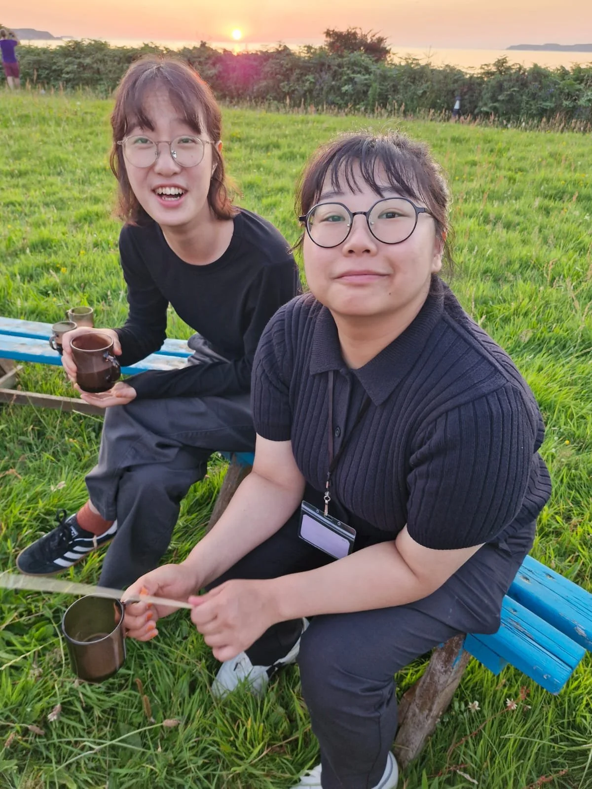 Two women sitting on a blue bench in a grassy field at sunset, holding brown cups, smiling at the camera, with a scenic background of water and trees.