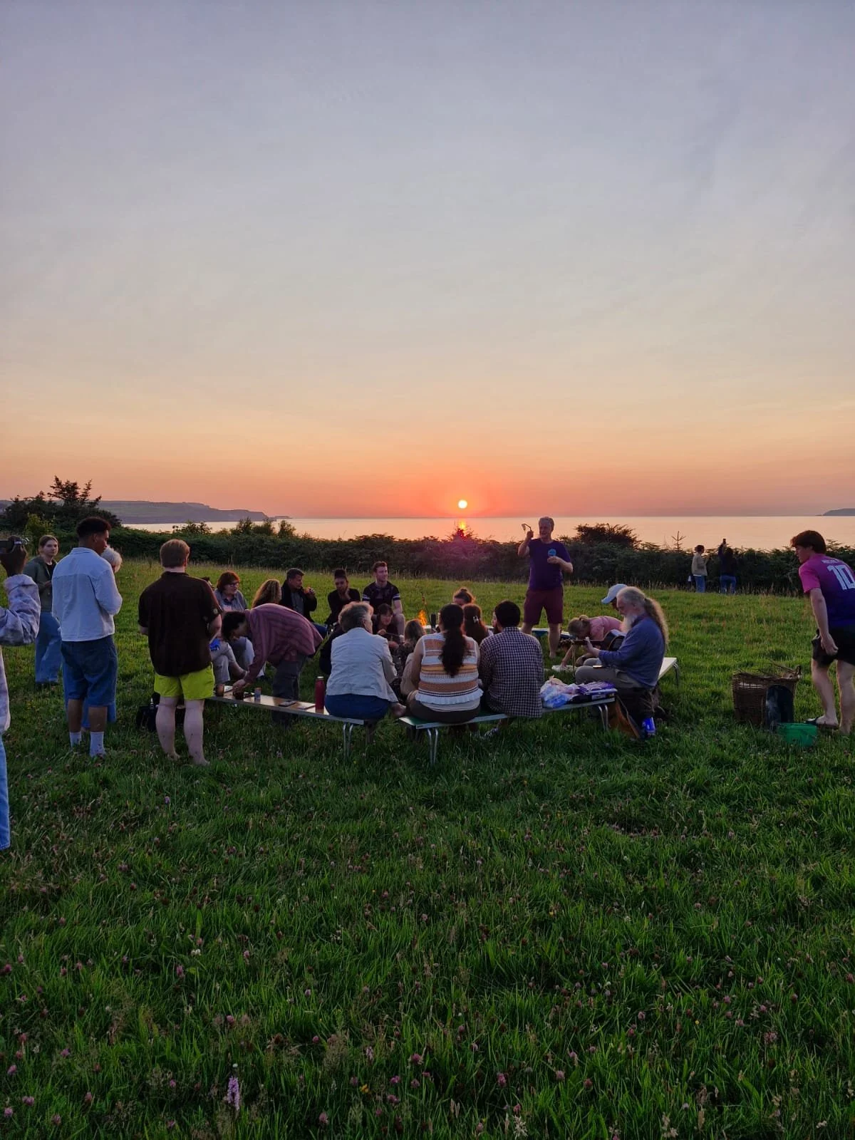 Group of people gathering outdoors on a grassy hill at sunset, sitting around a low table, with others standing nearby. The scene features a body of water and the sun setting on the horizon, creating a warm, colorful sky.