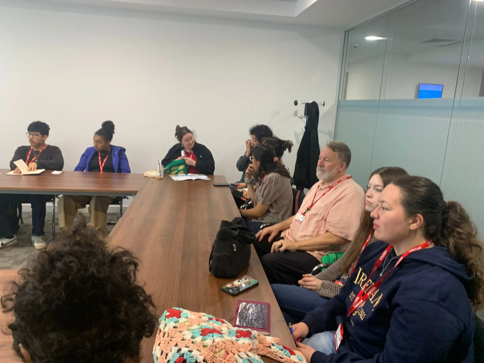 A group of diverse individuals, including young students and adults, sitting around a conference table in a modern room, some taking notes and others listening attentively.