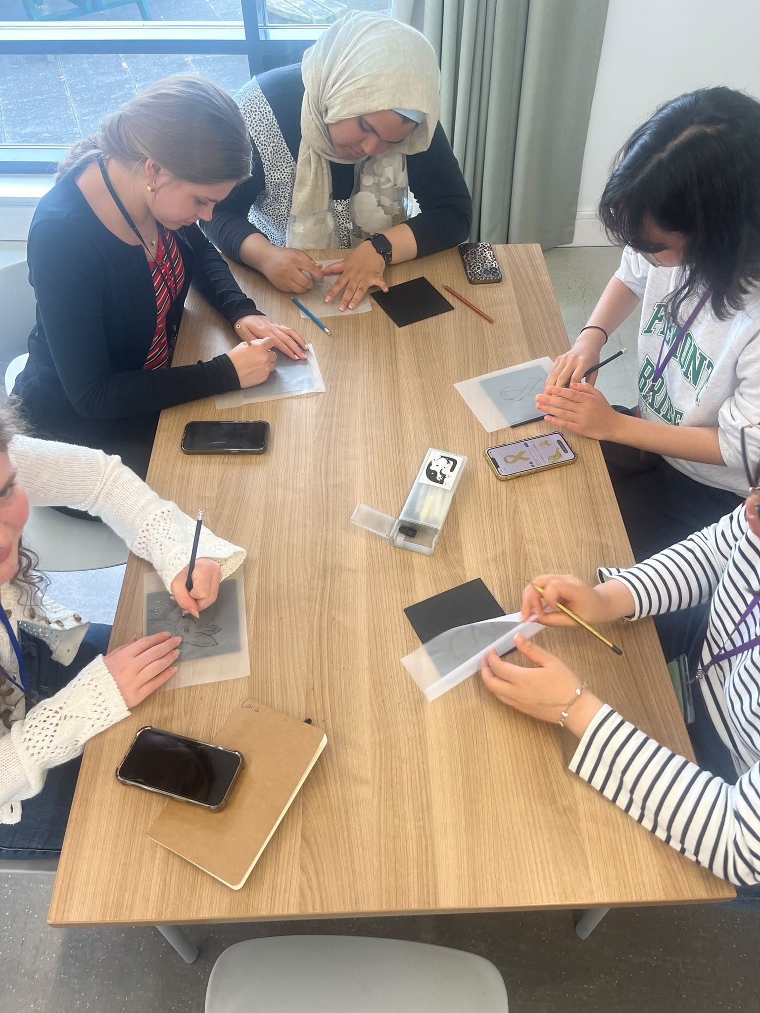 Several people sitting around a wooden table, working on small projects with black and white paper, pencils, and brushes. There are smartphones and a small container on the table, with a background of large windows and curtains.