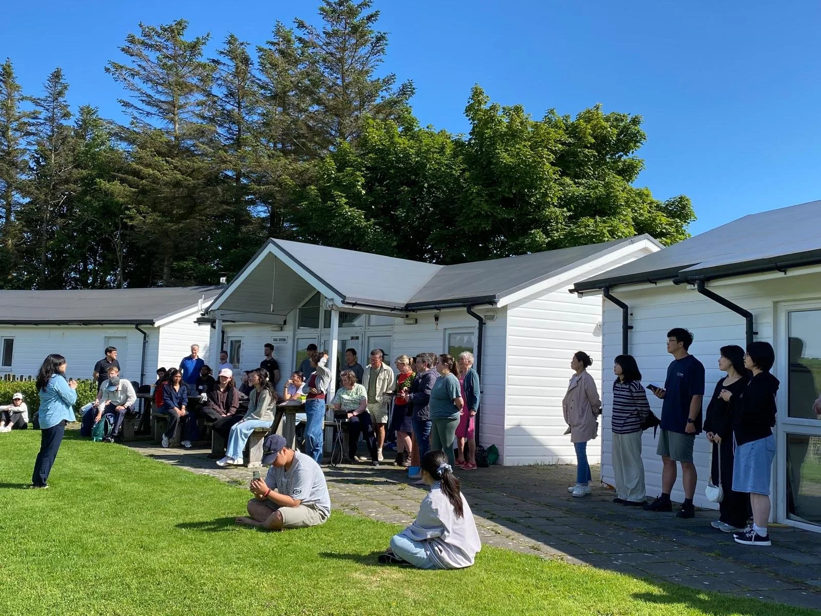 A large group of people gathered outdoors on a sunny day, standing and sitting on the lawn and patio in front of white houses with black gutters. Some are seated while others are standing and listening to a woman speaking.