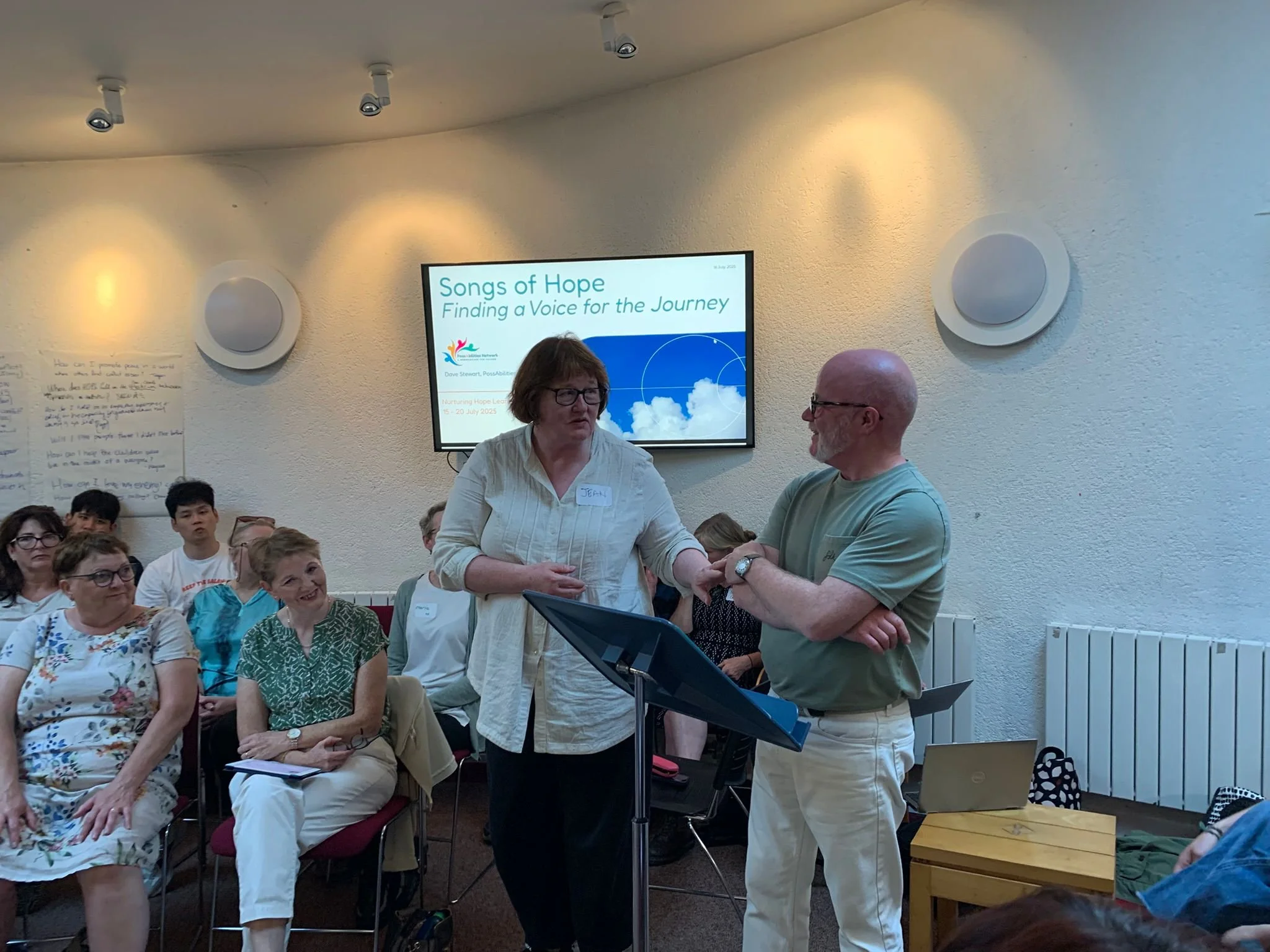 A woman and a man are engaged in a conversation during a presentation titled 'Songs of Hope: Finding a Voice for the Journey' in a room with seated audience members. A large screen displays the presentation title. The woman is speaking, wearing glass
