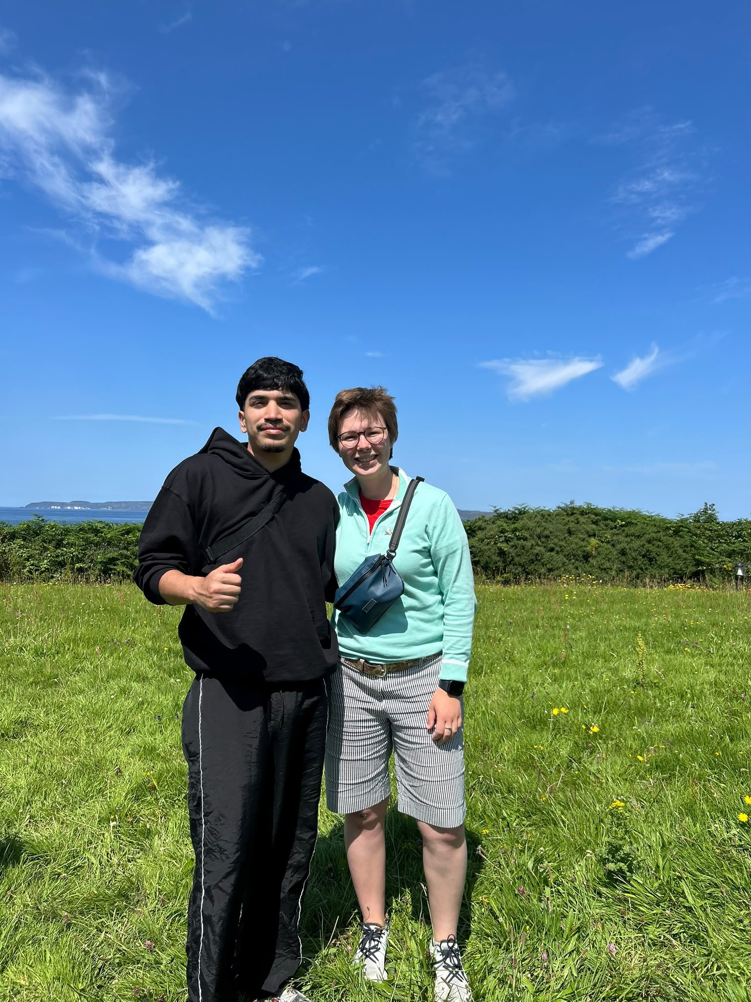 A man and a woman standing outdoors on a grassy field with trees, a body of water, and a blue sky with some clouds in the background.