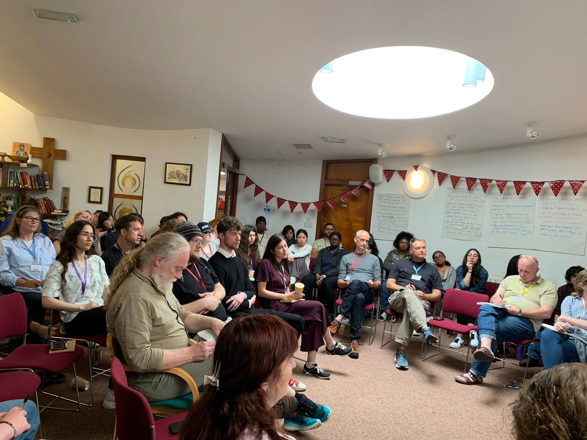 A diverse group of people sitting in a circle in a room, attending a workshop or seminar, with whiteboards on the wall and red bunting decorations.