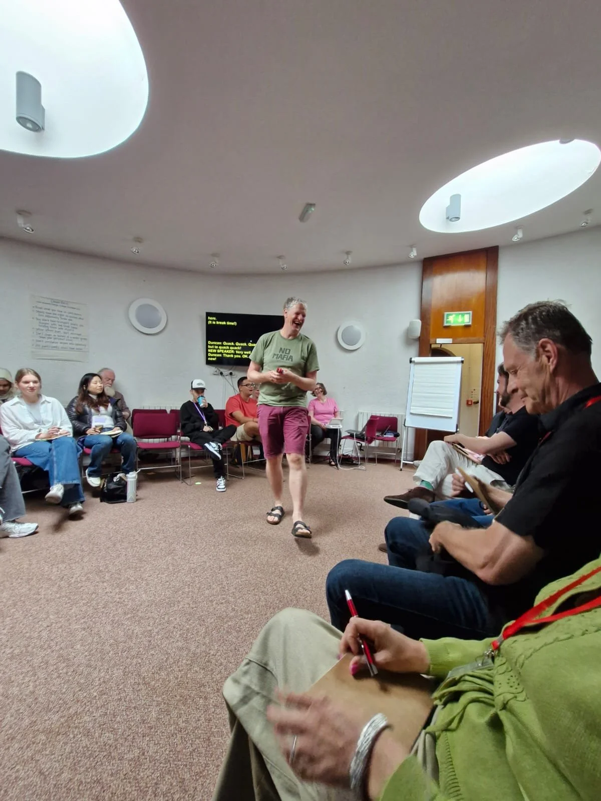 A group of people seated in a circle in a conference room. A man in shorts and sandals is walking and smiling at the center, while others are watching and taking notes.