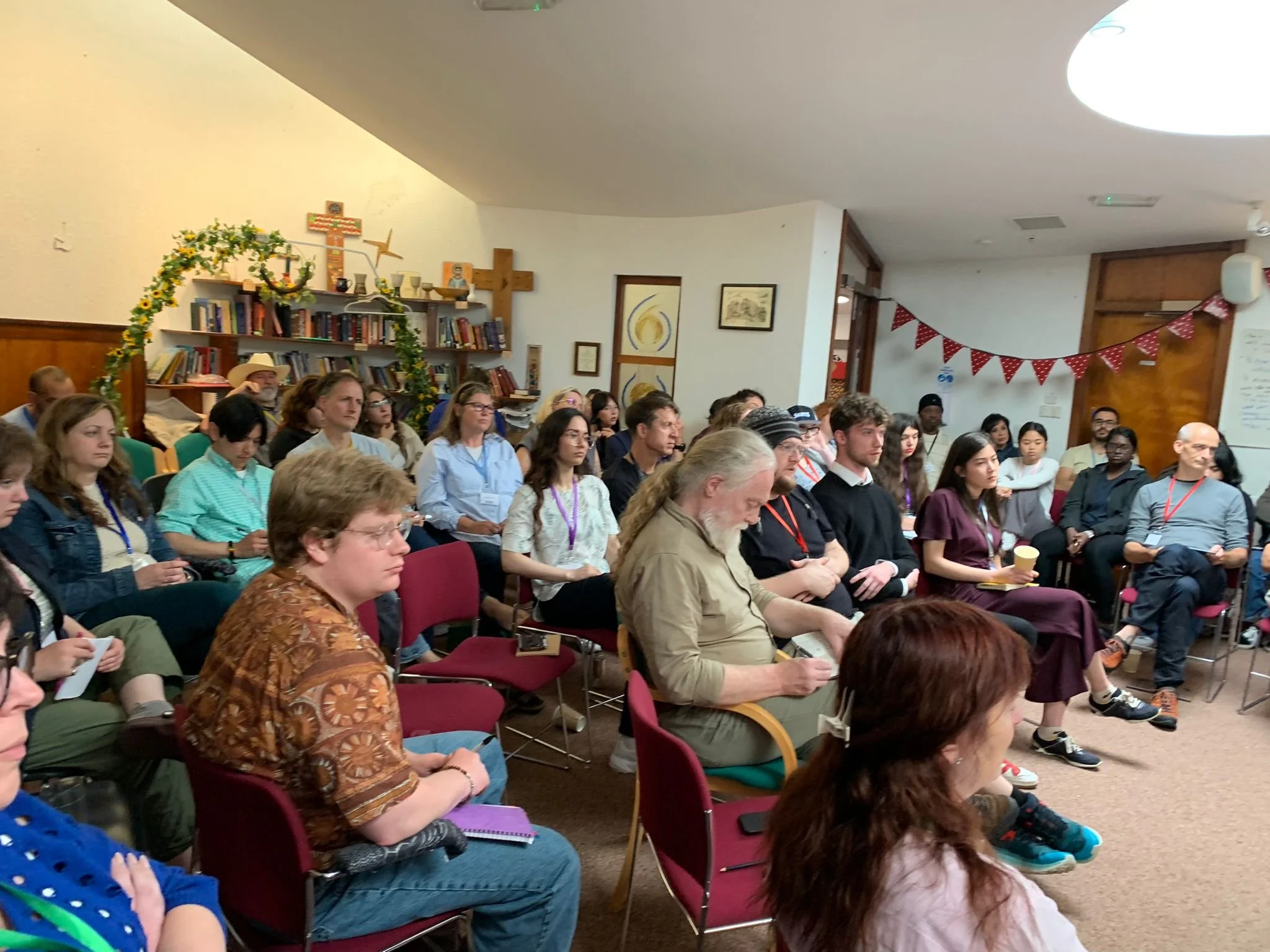 A group of people sitting attentively in a room with bookshelves and religious decorations, including crosses, and red and white bunting.