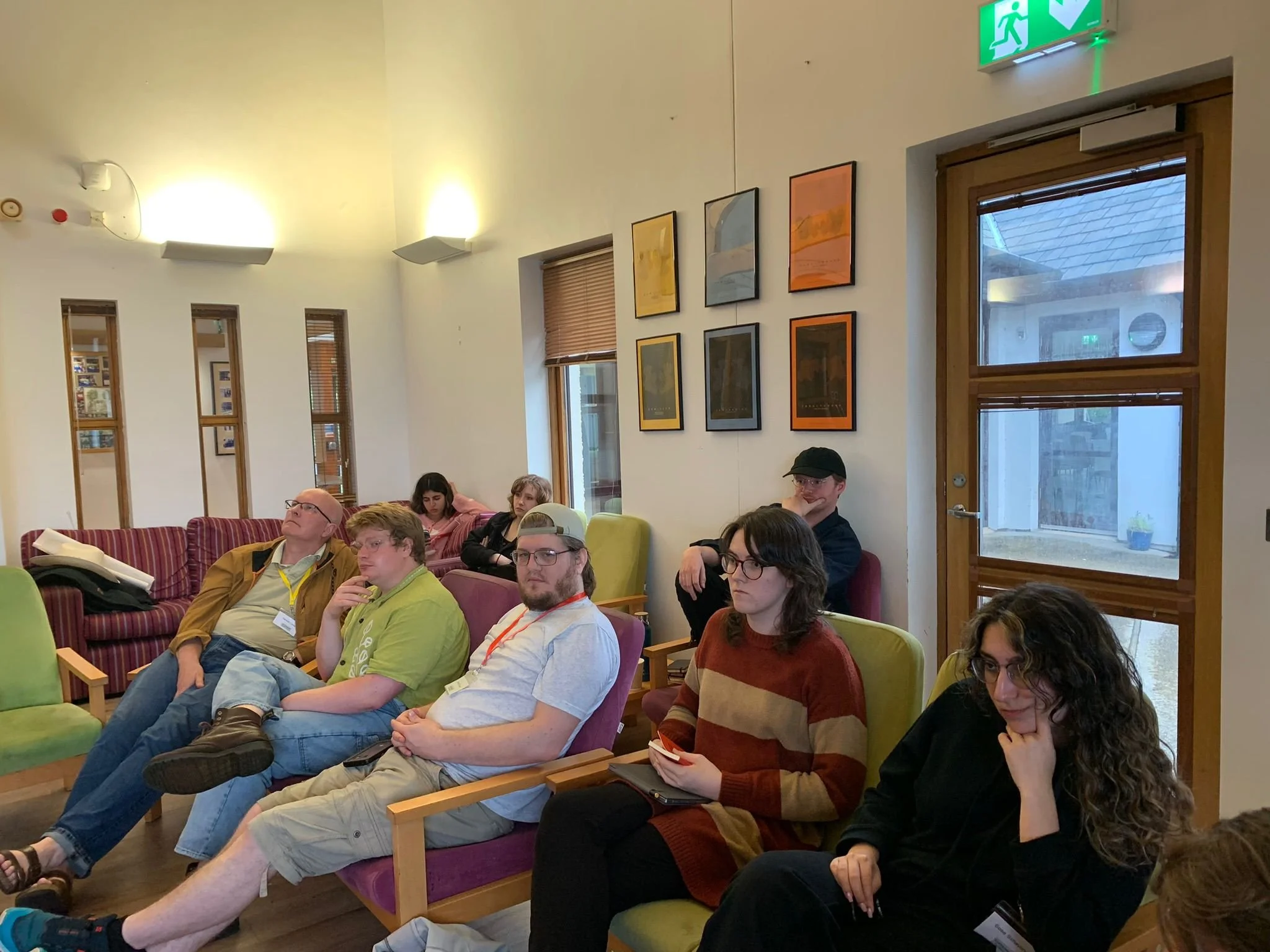 Group of people sitting on chairs and sofas in a room, some taking notes, appearing attentive during a presentation or discussion.