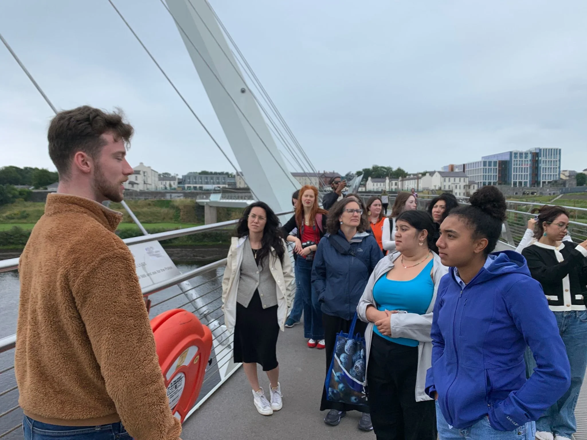 A group of people listening to a tour guide on a bridge with modern buildings and a river in the background.