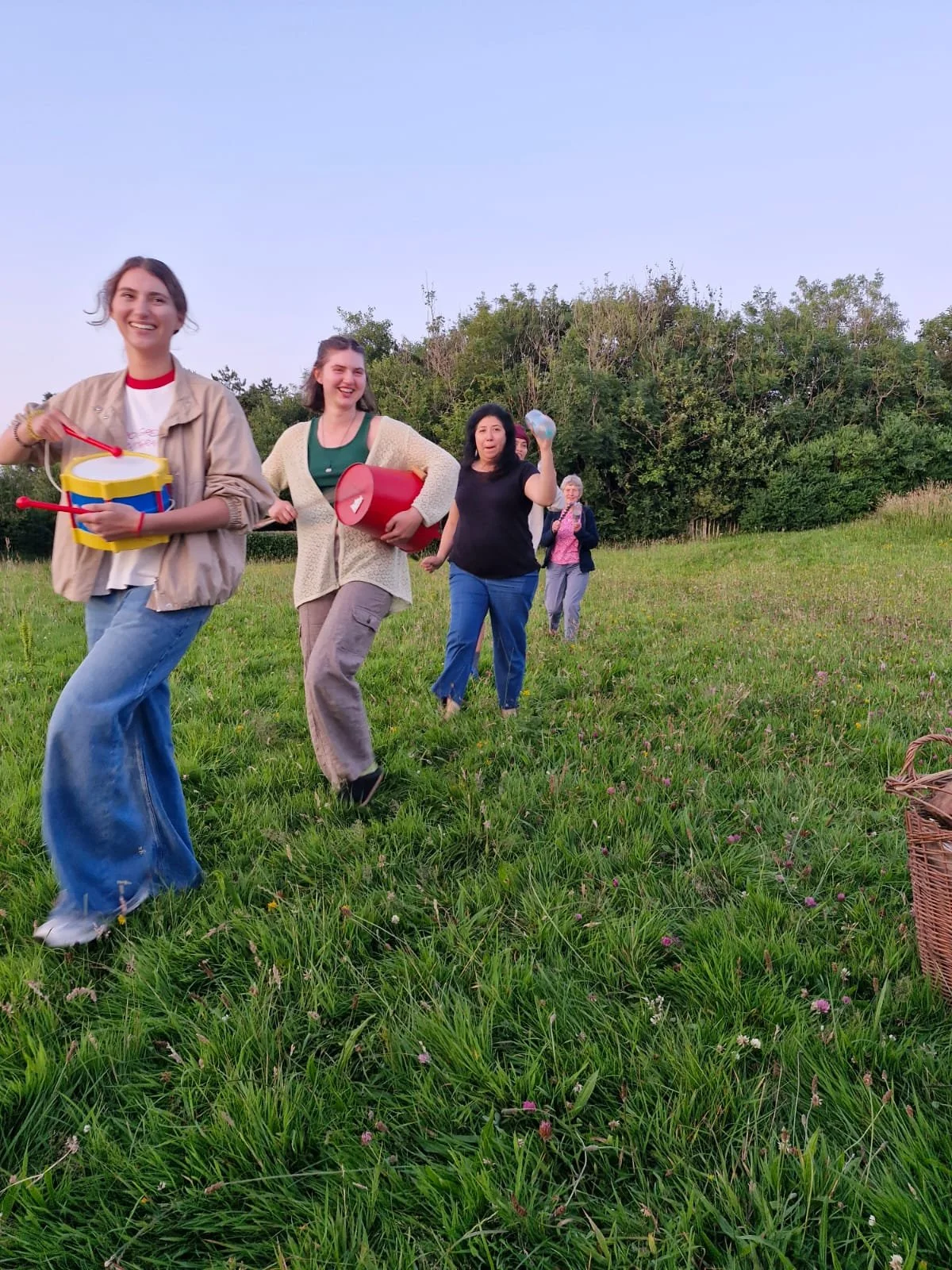 Group of women walking through a grassy field, smiling, holding various objects, with trees in the background.