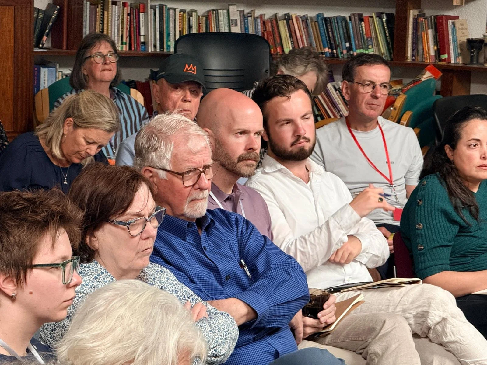 A group of people sitting in an indoor setting, attentively listening. Background shows a bookshelf filled with books.