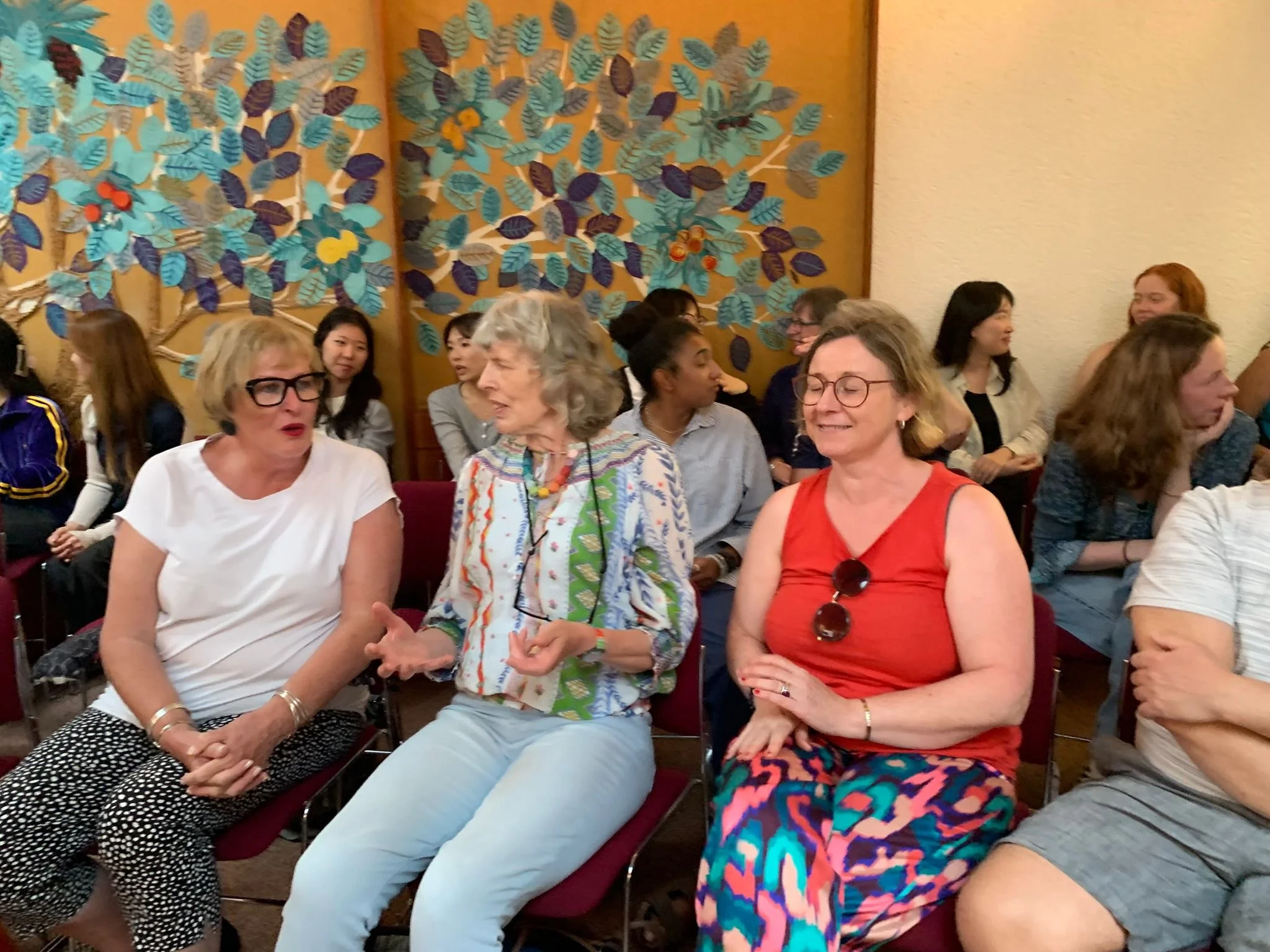 A group of women sitting and talking in a room with a colorful leaf-themed mural on the wall.