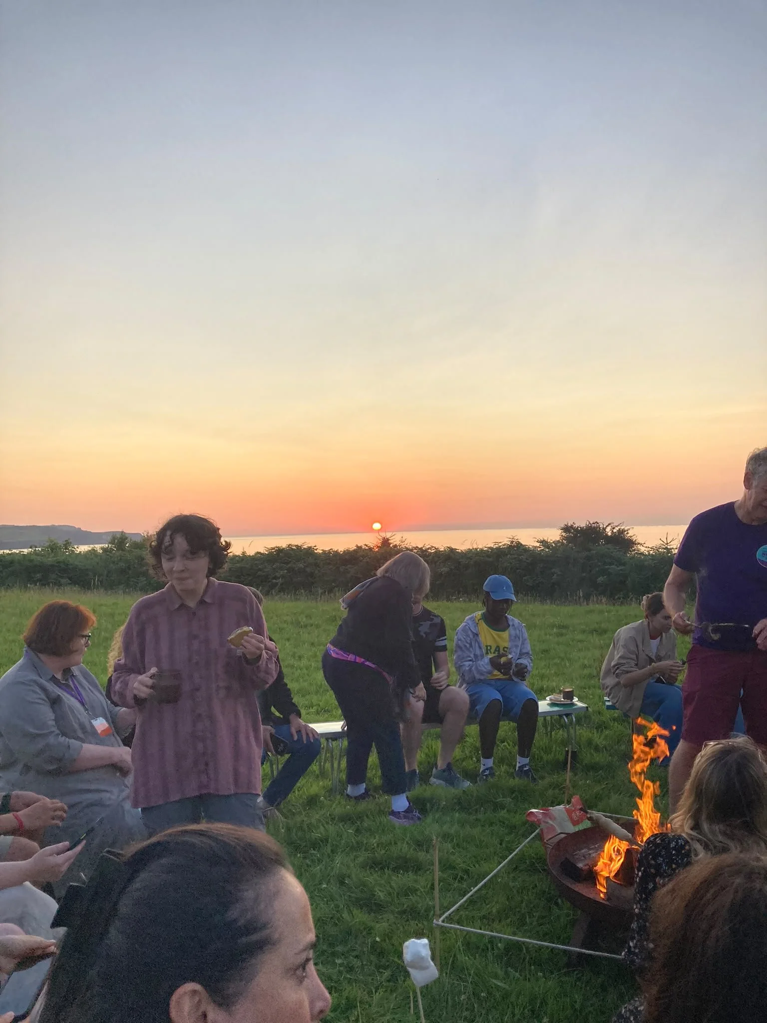 People gathered outdoors around a fire pit during sunset, some sitting on chairs and some standing, with greenery and a sunset sky in the background.
