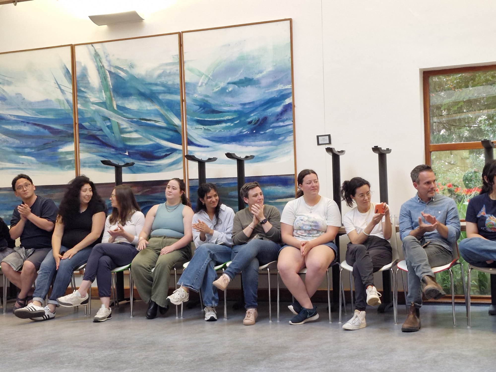 People sitting in chairs in a room, some clapping, others taking photos, with large windows and a blue abstract painting on the wall.