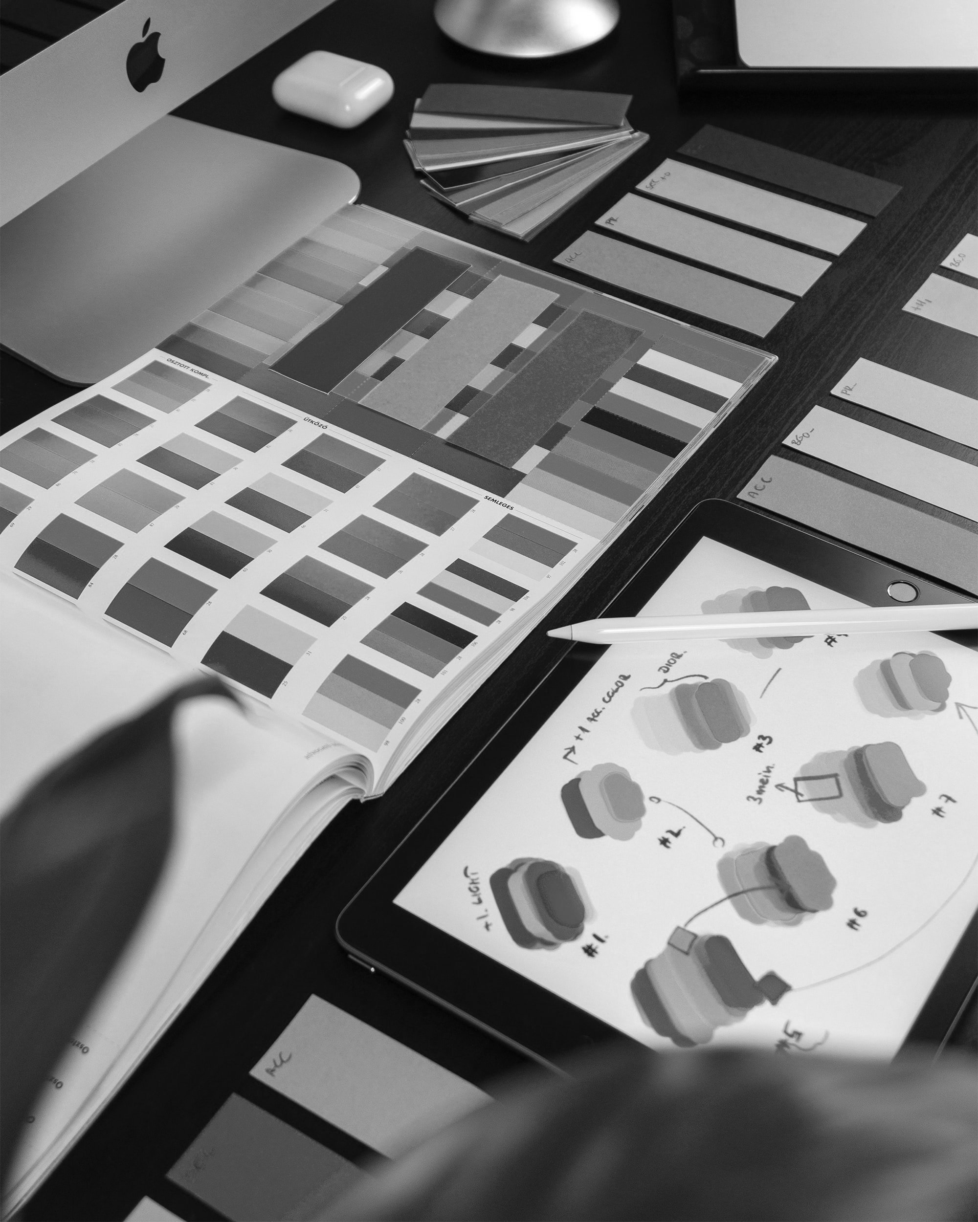 A workspace with color swatches, fabric samples, and design sketches, including a digital tablet with a 3D color palette and a pen, surrounded by color labels and a laptop.