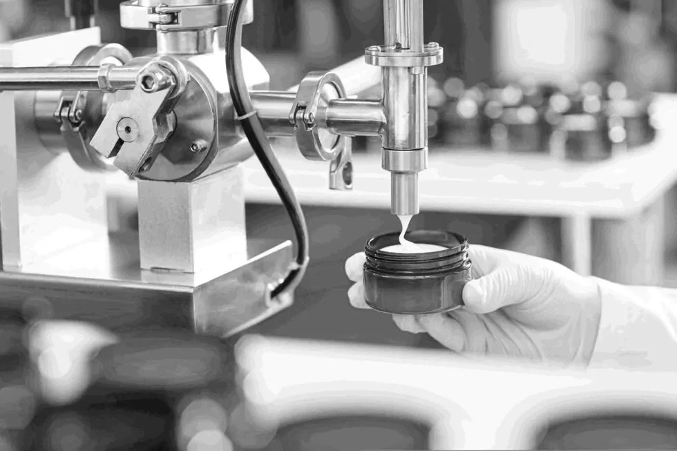 A person wearing gloves is filling a small jar with a creamy substance from a filling machine in a laboratory or industrial setting.