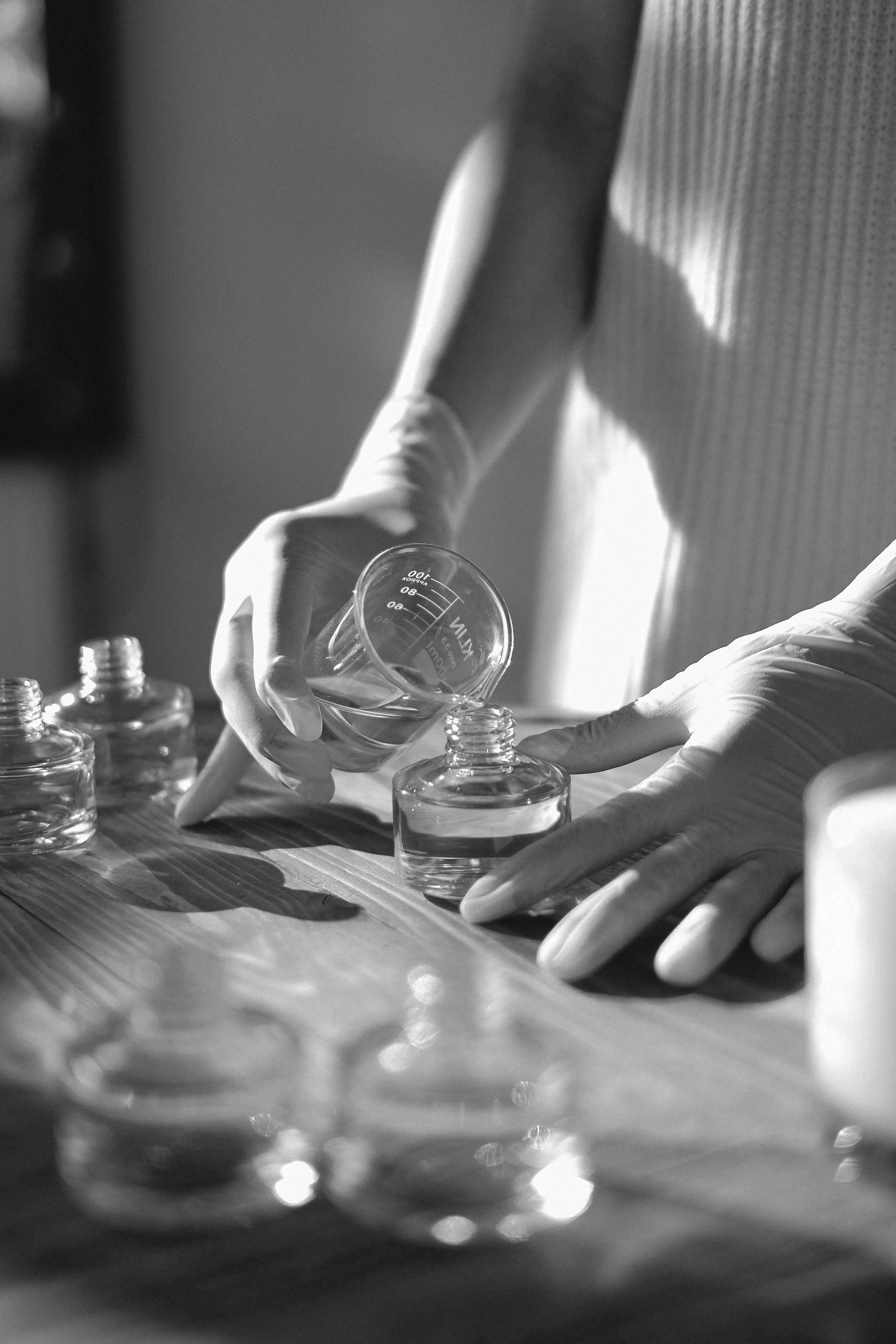 Person wearing gloves pouring a liquid from a beaker into a small glass container on a wooden table, with other glass containers nearby, in black and white.