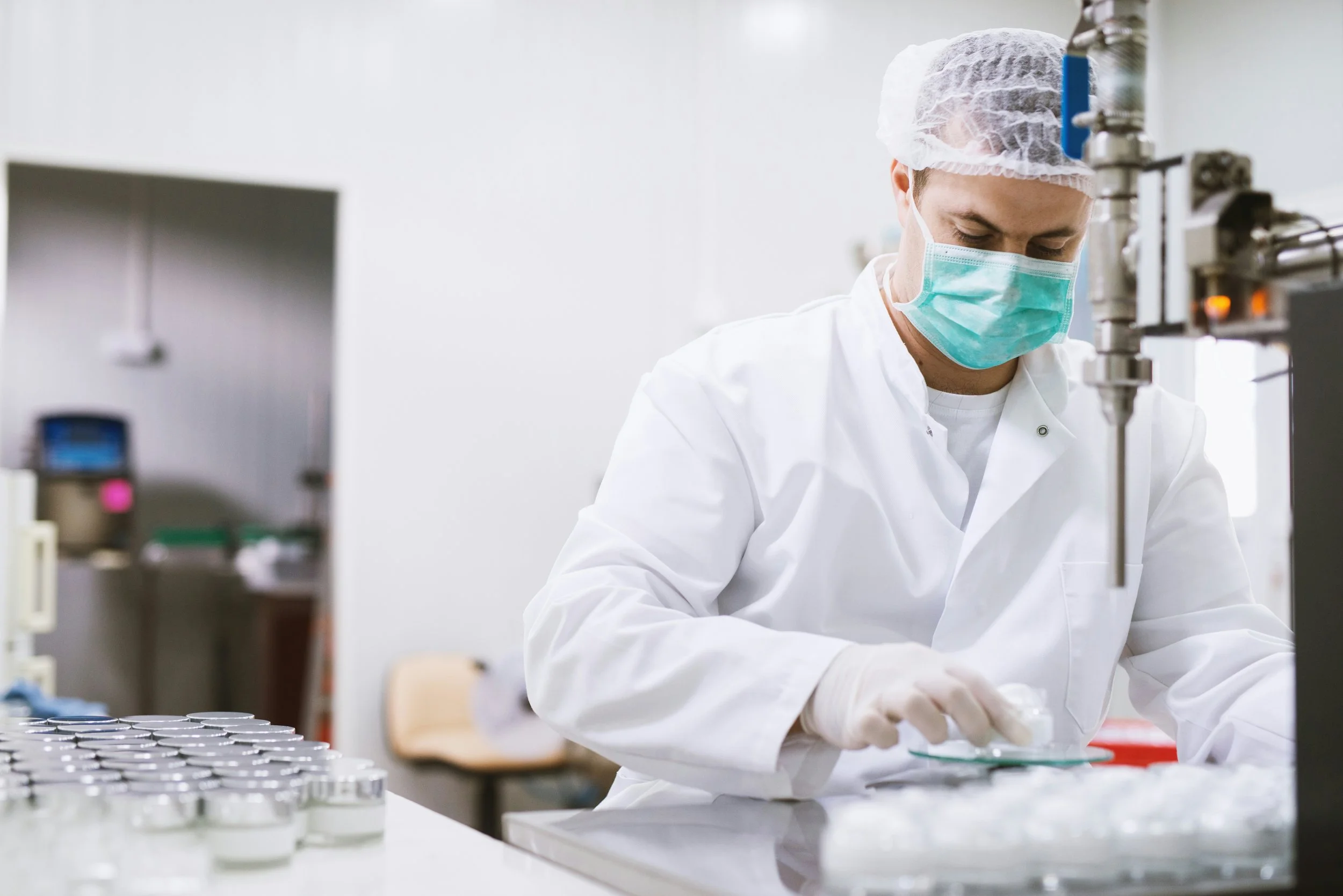A scientist in a lab, wearing a white lab coat, blue surgical mask, gloves, and a hairnet, working with scientific equipment.