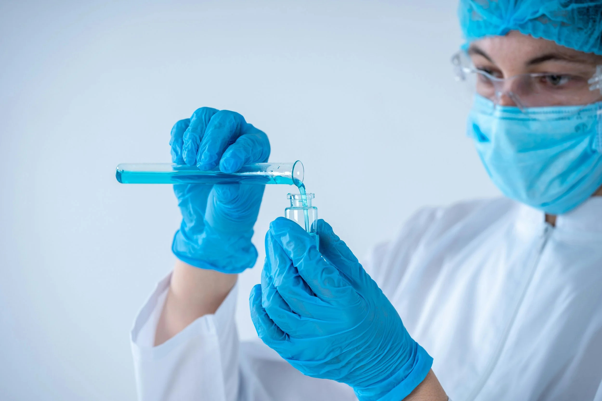 A scientist wearing a blue face mask, safety glasses, and blue gloves is pouring a blue liquid from a test tube into a small beaker.