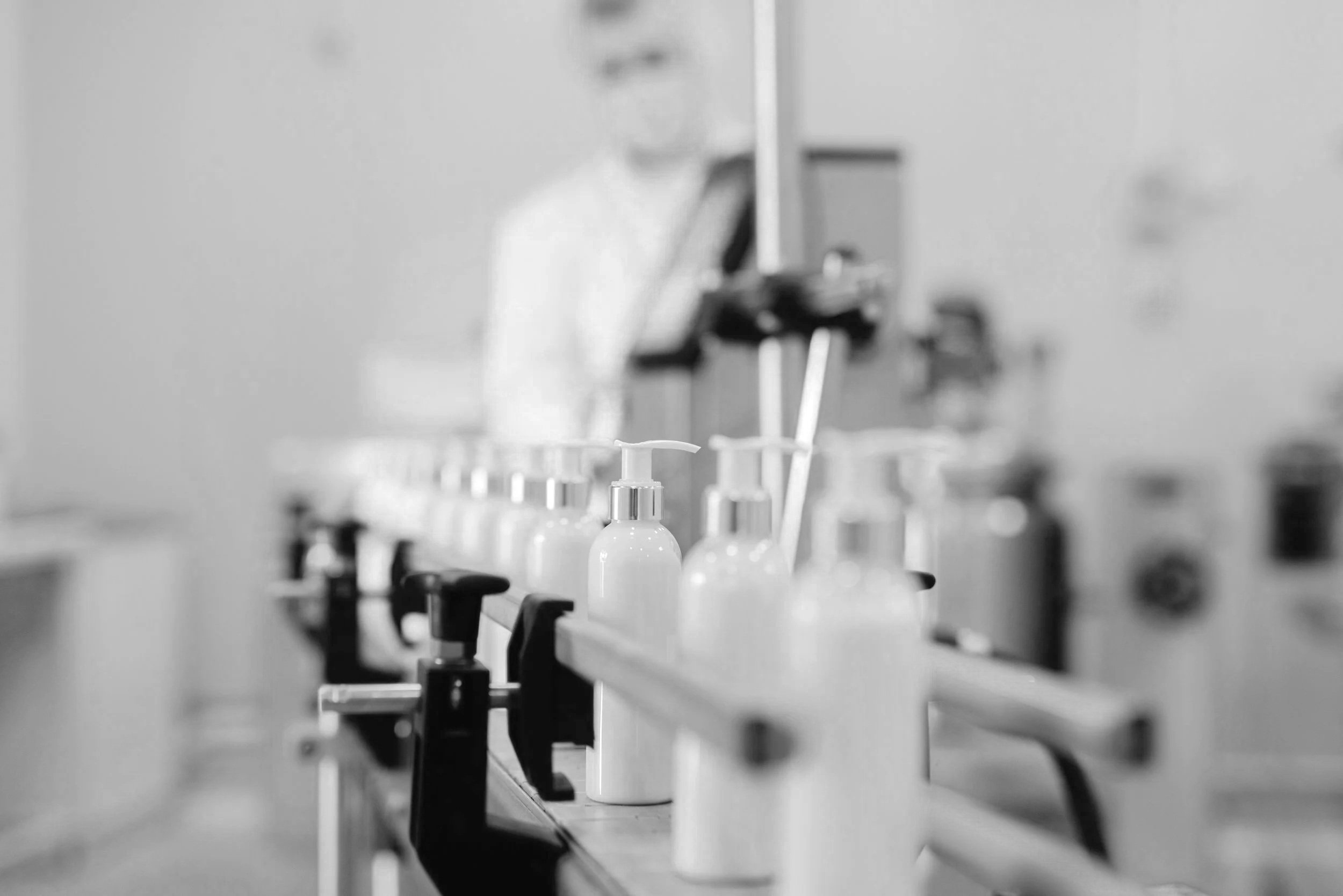 Black and white photo of a laboratory or medical setting with row of bottles on a laboratory bench and a blurred person in the background.